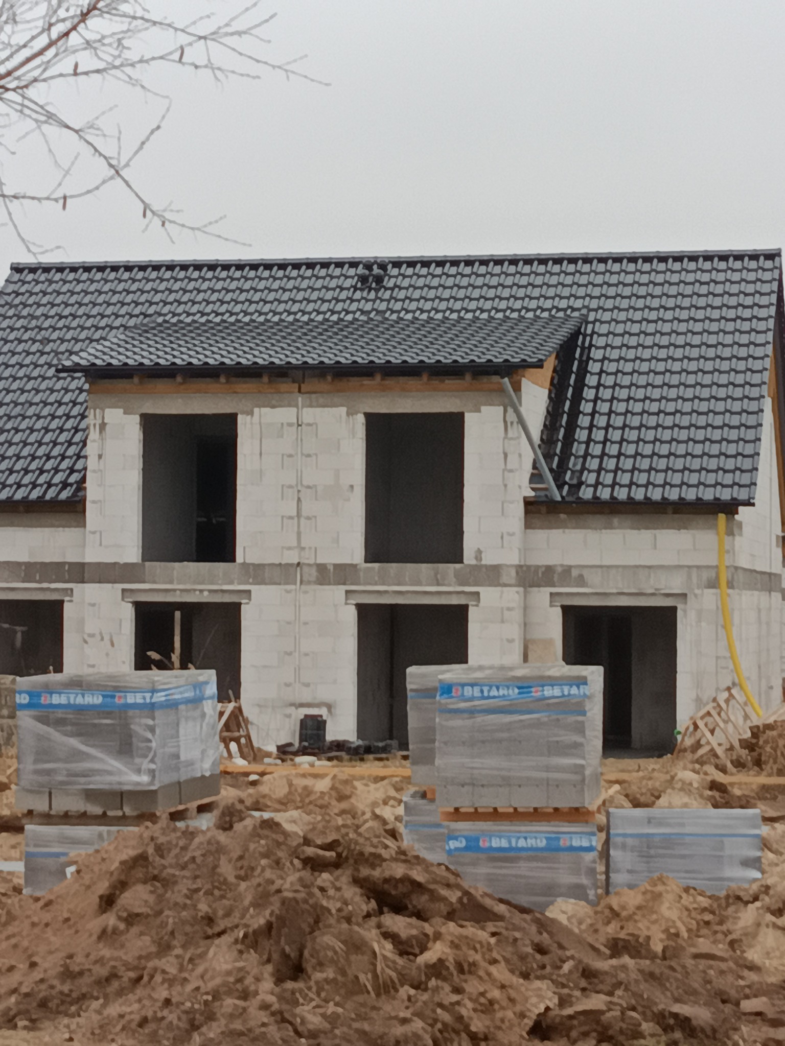 Surowy stan domu z bloczków, czarny dach z rynną, palety z materiałem budowlanym na pierwszym planie, ziemia, pochmurne niebo.