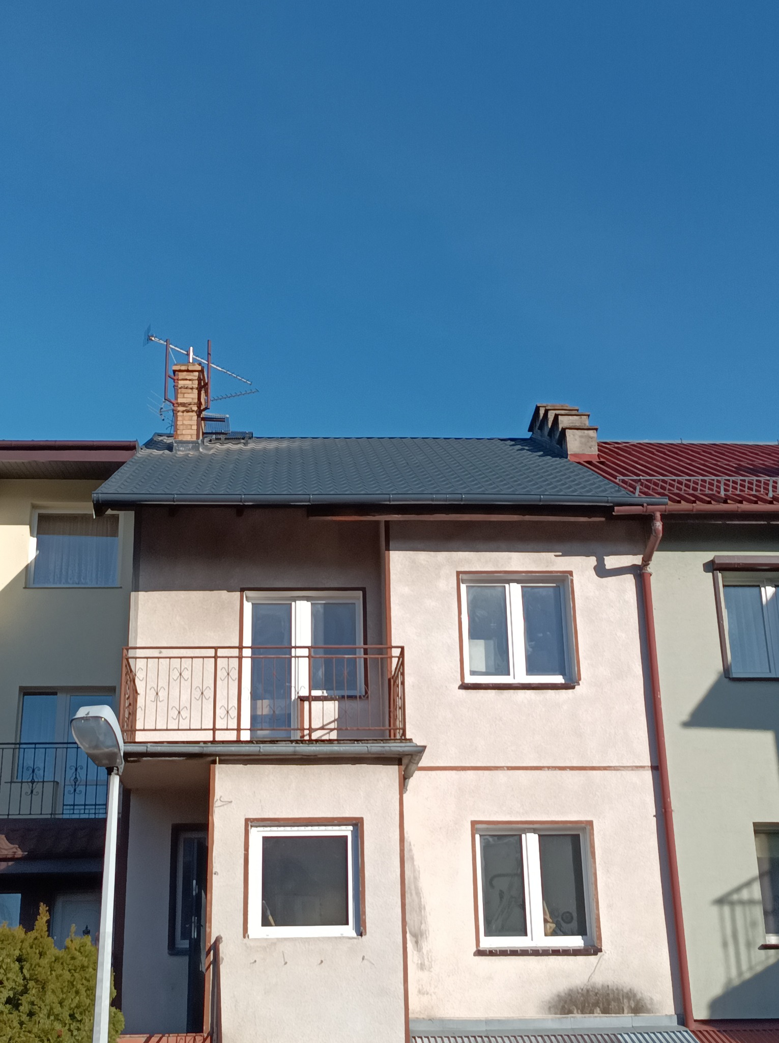 Szary dach z blachy na dwupiętrowym budynku z balkonem, widoczny komin z anteną i fragment dachu sąsiedniego budynku z czerwoną dachówką na tle błękitnego nieba.