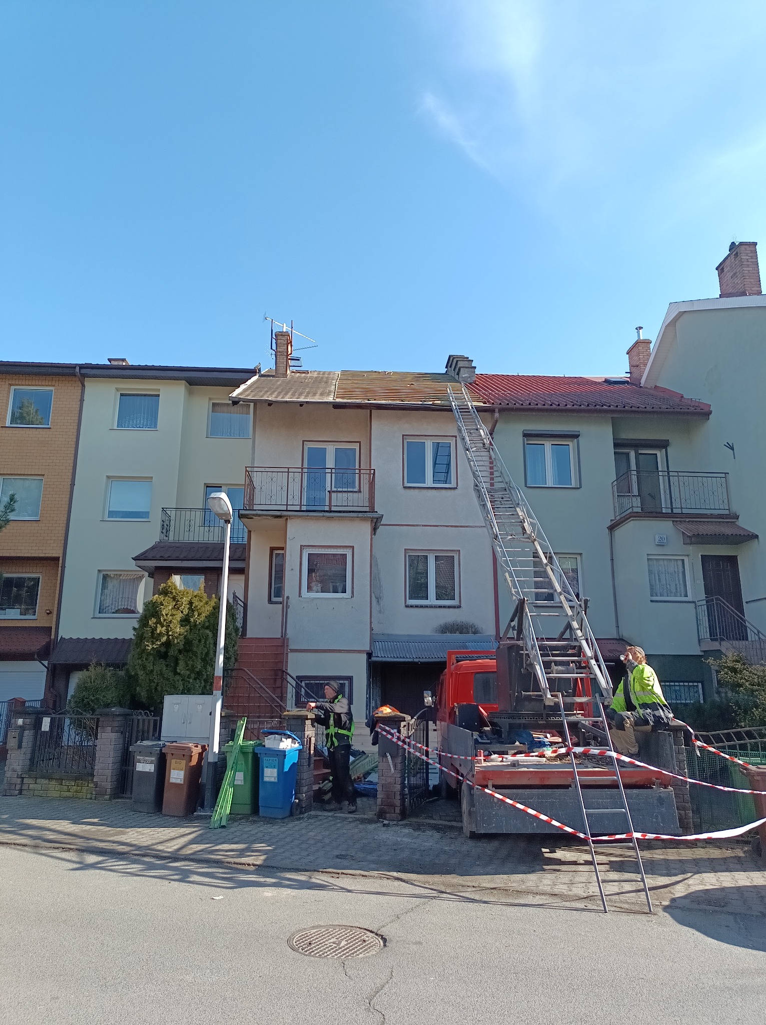 Ekipa na drabinie przy dachu budynku podczas prac. Widoczny samochód z drabiną, pracownicy w strojach roboczych i zabezpieczenia. Częściowo zdjęty dach.