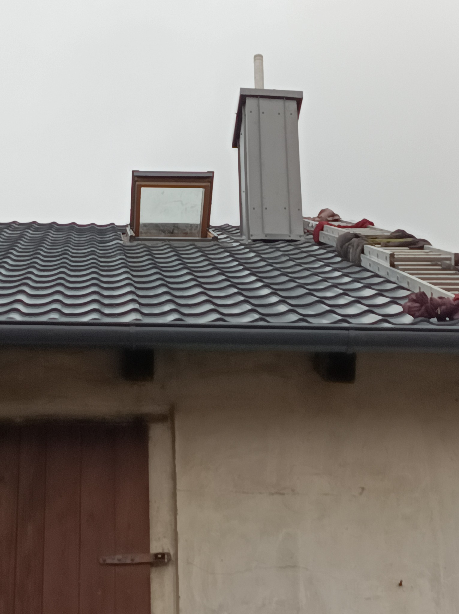 Dach z blachodachówki z oknem dachowym i kominem. Na dachu drabina dekarska. Widoczny fragment ściany budynku z drewnianymi drzwiami.