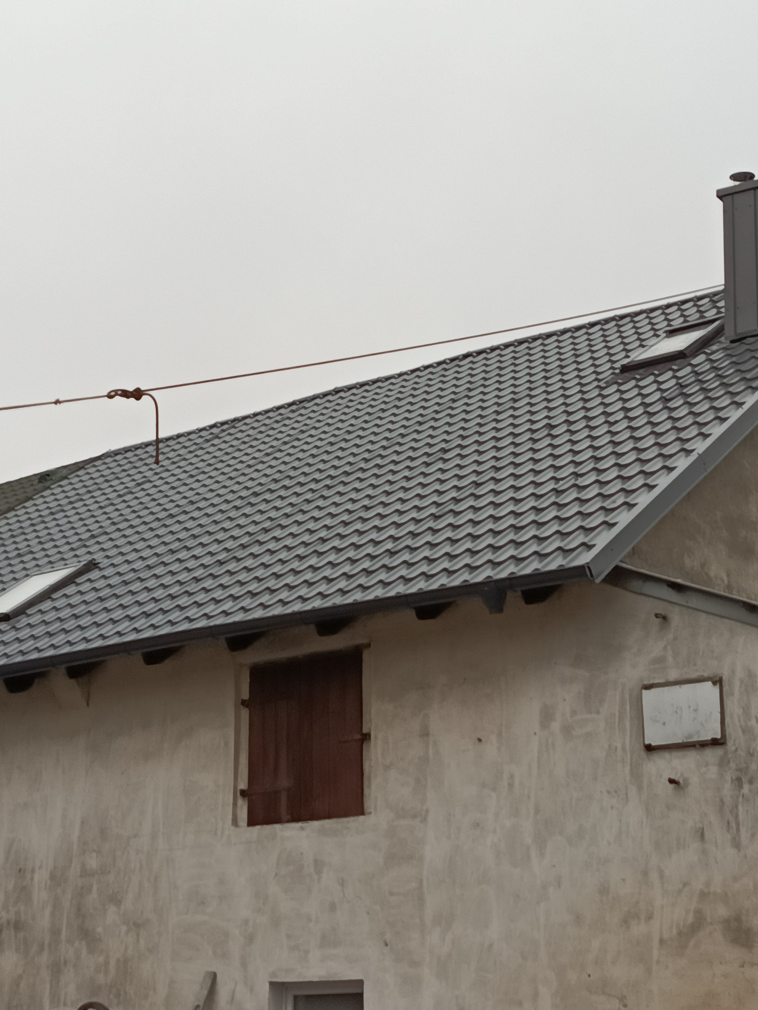 Szary dach z dachówki ceramicznej na budynku z oknem z drewnianymi okiennicami. Widoczny komin i instalacja odgromowa. Tynk z widoczną fakturą.