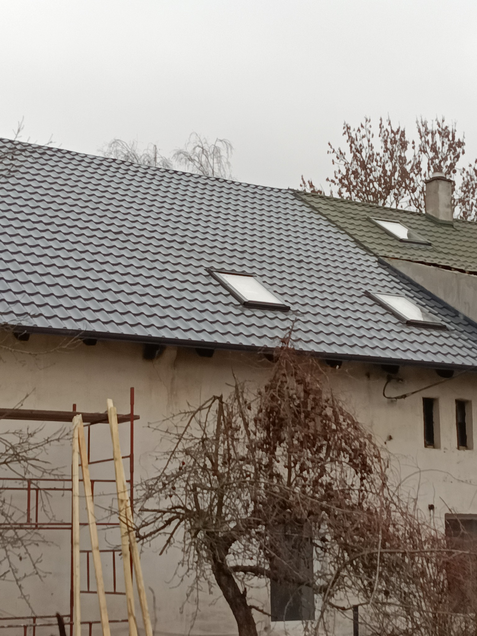 Szary dach z oknami dachowymi na białym budynku, widoczne rusztowania i drzewo bez liści. Część dachu ma inny, zielonkawy odcień.