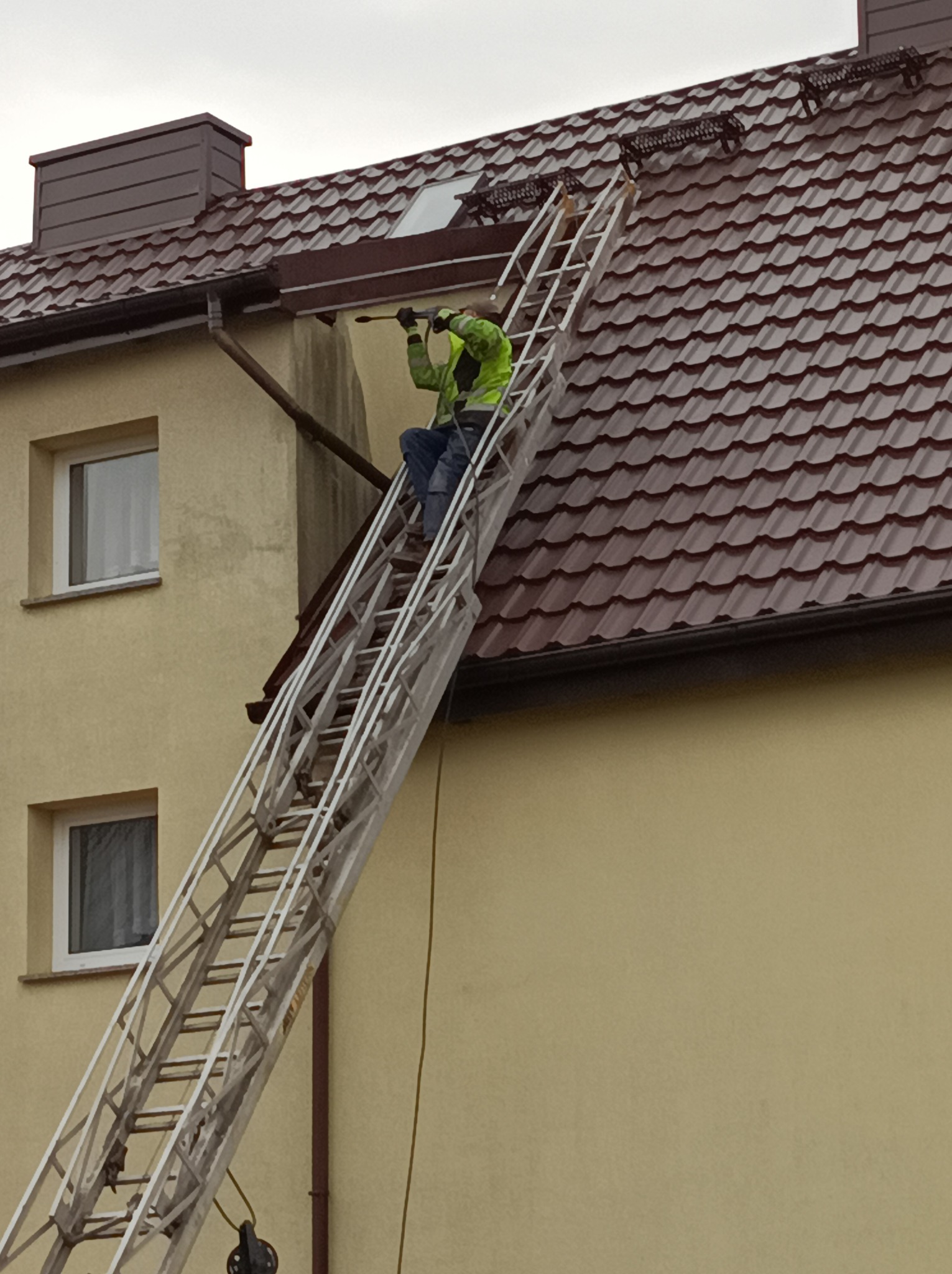 Pracownik w zielonej kurtce na drabinie czyści dach domu z brązową dachówką za pomocą myjki ciśnieniowej. Widoczne zabrudzenia na elewacji.