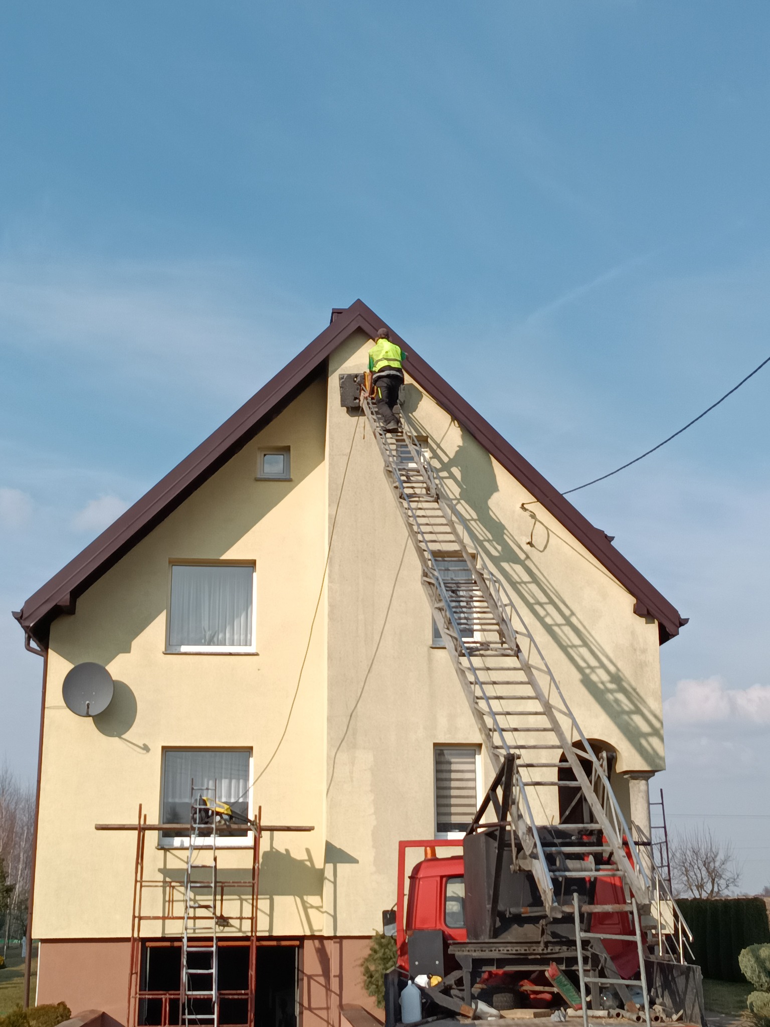 Pracownik na drabinie przy ścianie domu z brązowym dachem, prawdopodobnie wykonuje prace dekarskie. Widoczne rusztowanie i samochód z narzędziami.