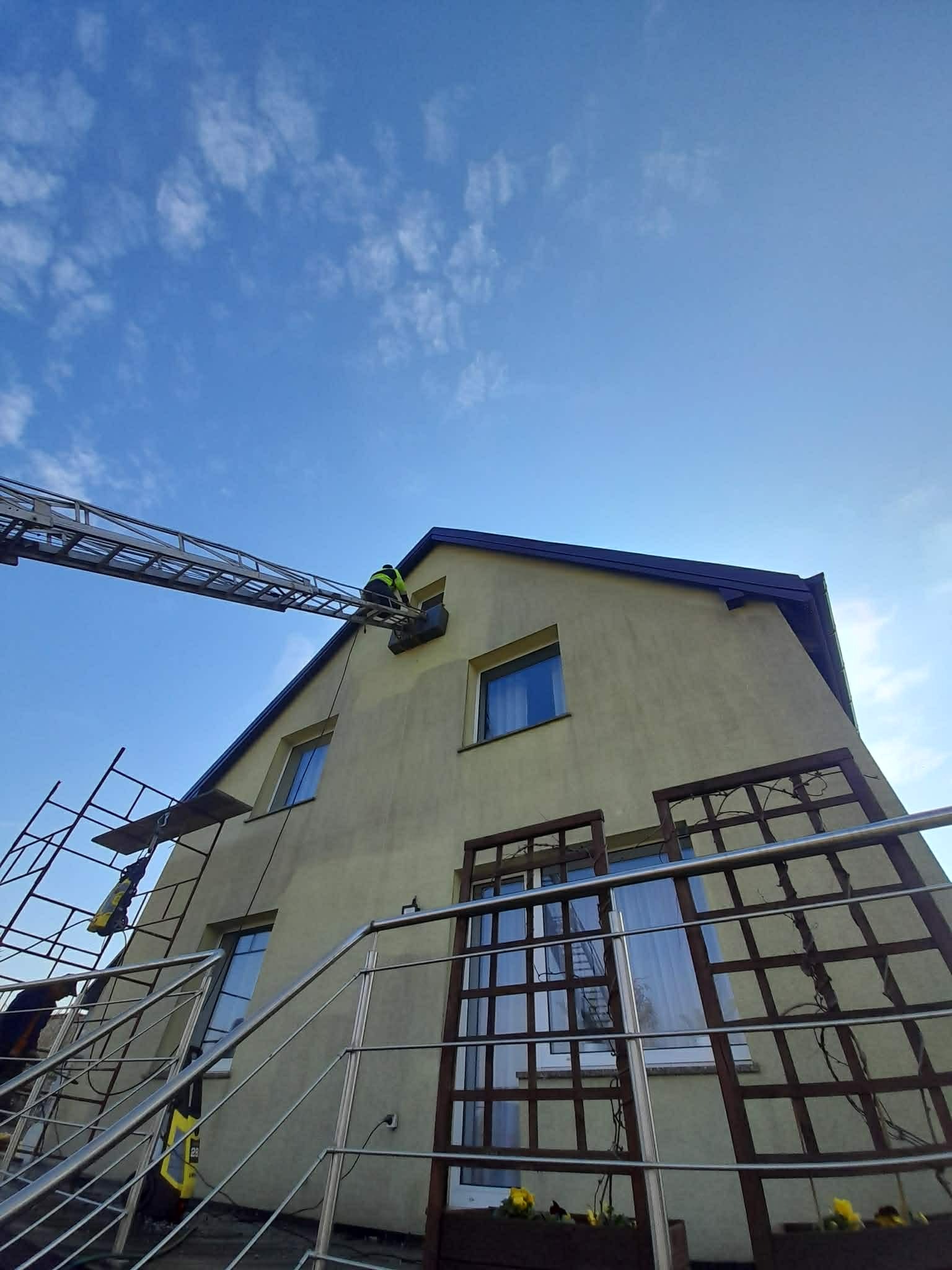 Pracownik na drabinie przy dachu domu z żółtą elewacją. Widoczne rusztowanie i błękitne niebo. Ujęcie z dołu, podkreślające wysokość budynku.