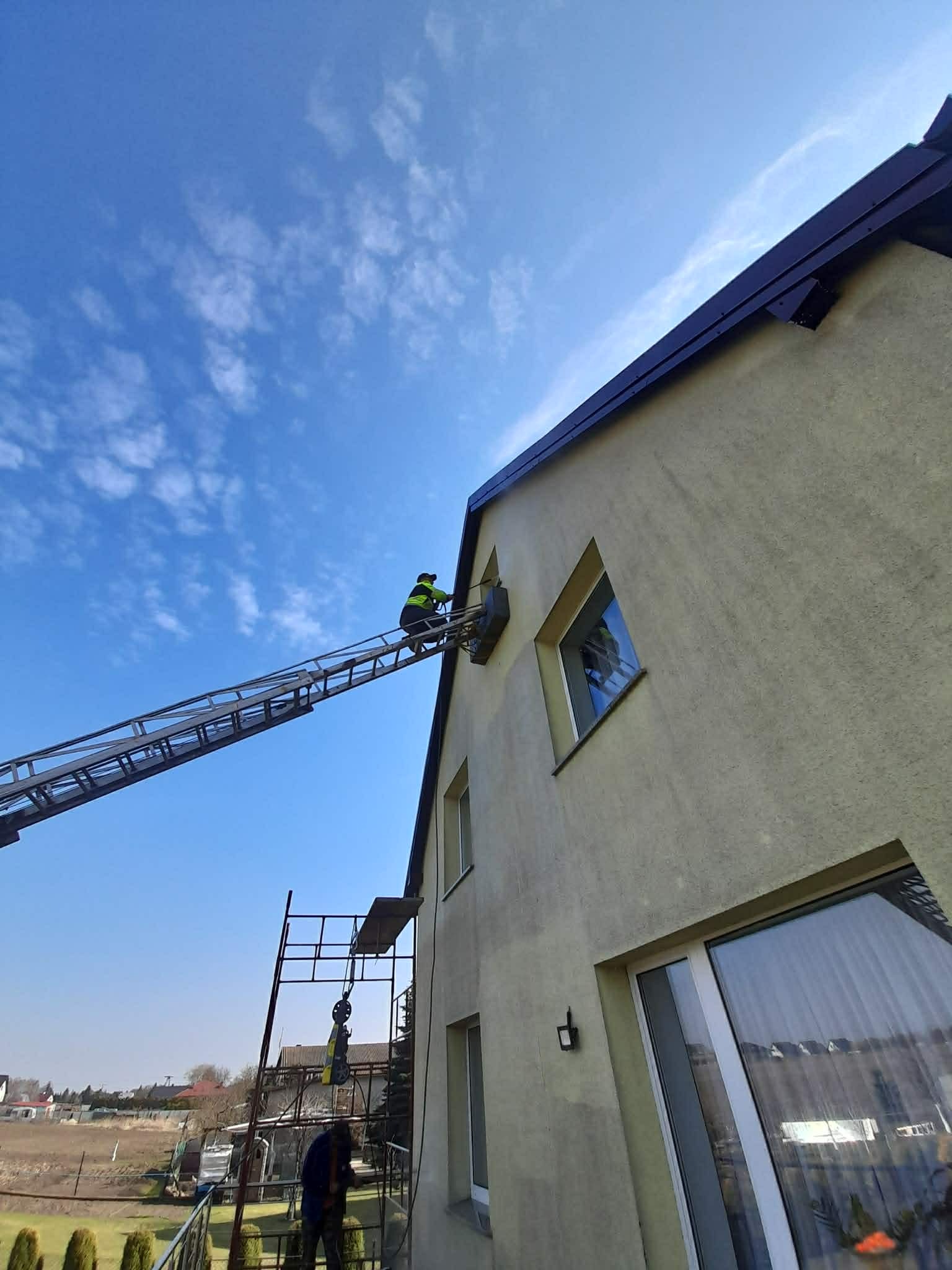 Pracownik na drabinie przy elewacji domu, prawdopodobnie montaż klimatyzacji. Widoczne rusztowanie i błękitne niebo.