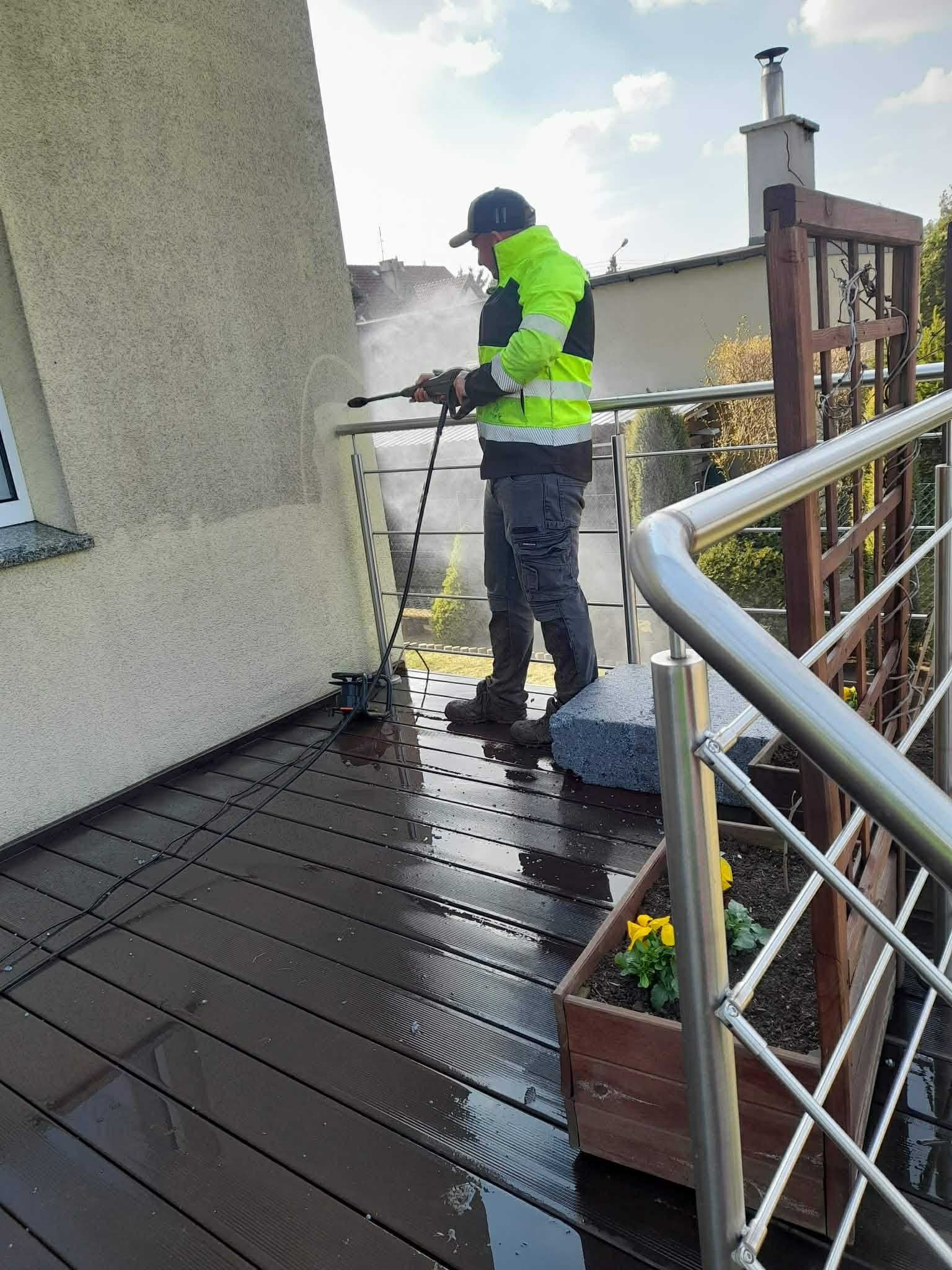 Mężczyzna w odblaskowej kurtce czyści elewację domu myjką ciśnieniową z balkonu. Widoczne zacieki i czysta powierzchnia. W tle fragment dachu i niebo.