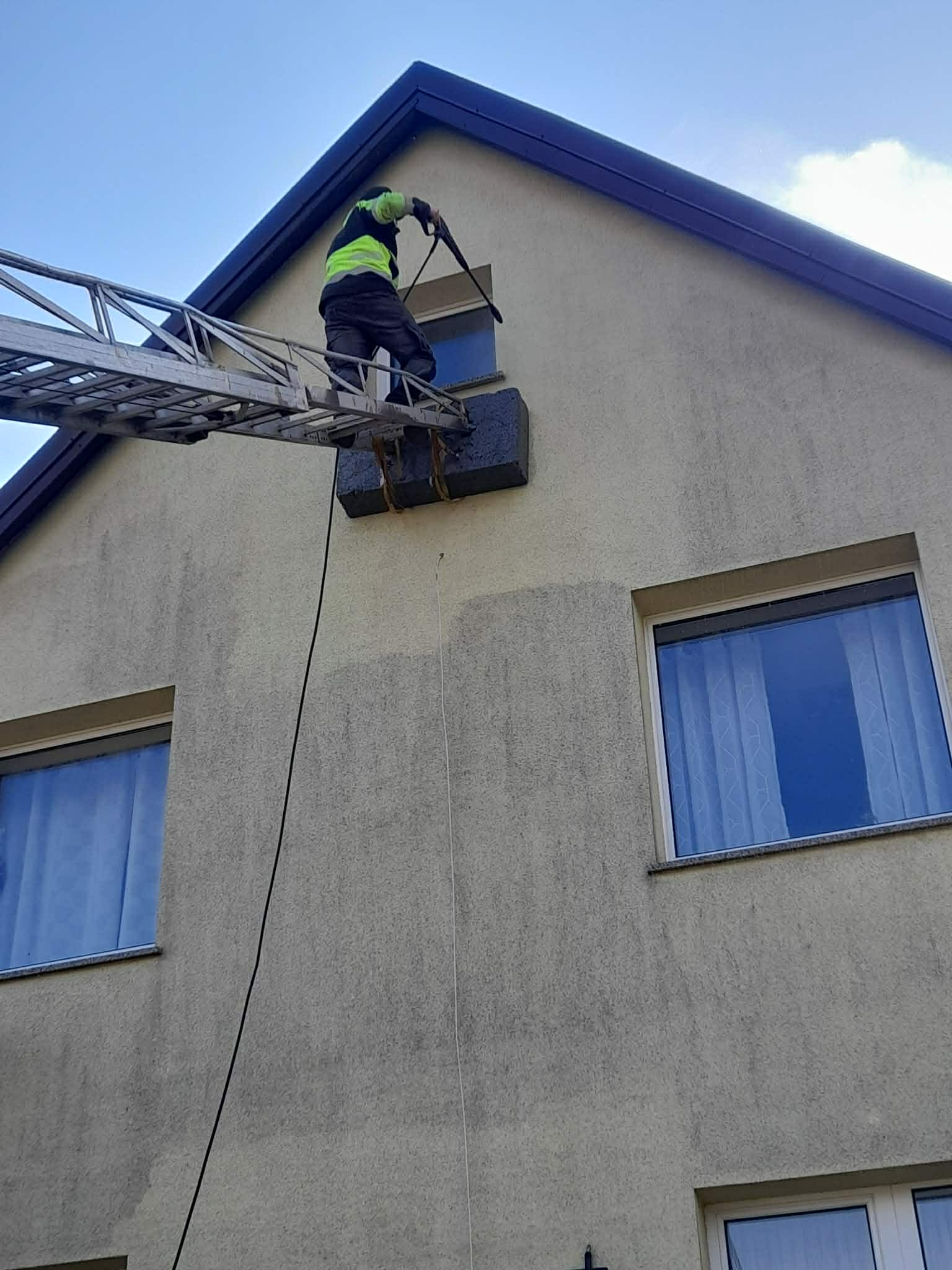 Pracownik w kamizelce na drabinie myje elewację domu. Widoczne okna i fragment dachu. Ujęcie z dołu, na tle błękitnego nieba.