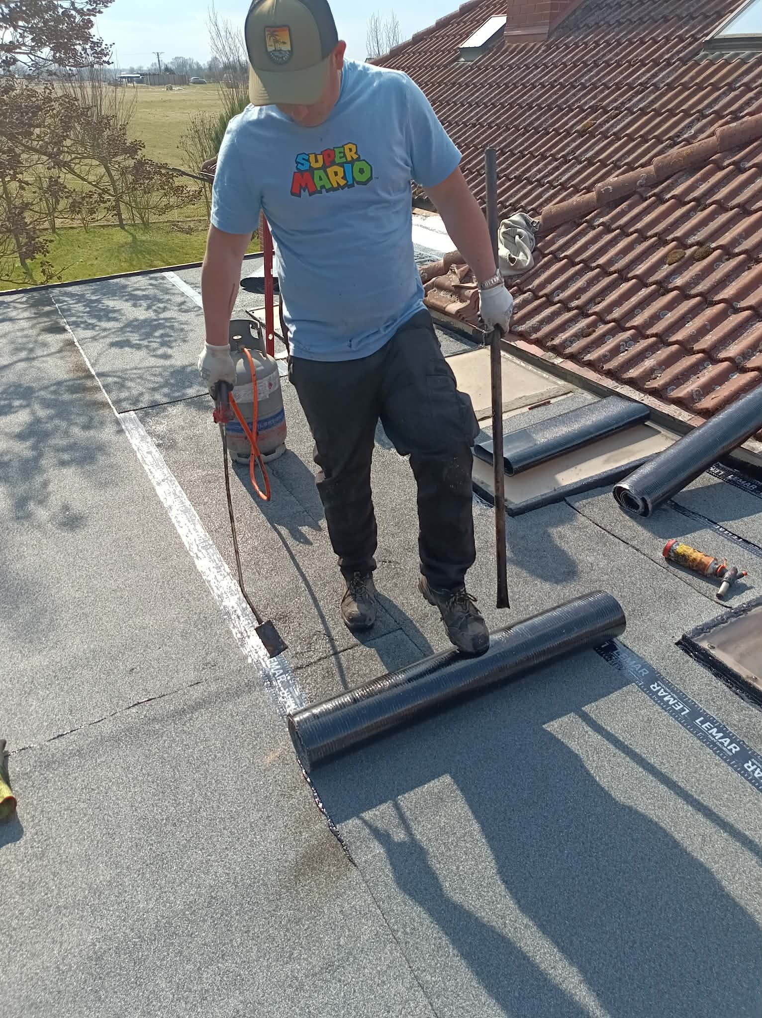 Praca na dachu: dekarz z palnikiem i rolką papy termozgrzewalnej na płaskim dachu. Widoczne narzędzia i fragment dachu z dachówką w tle. Dzień słoneczny.