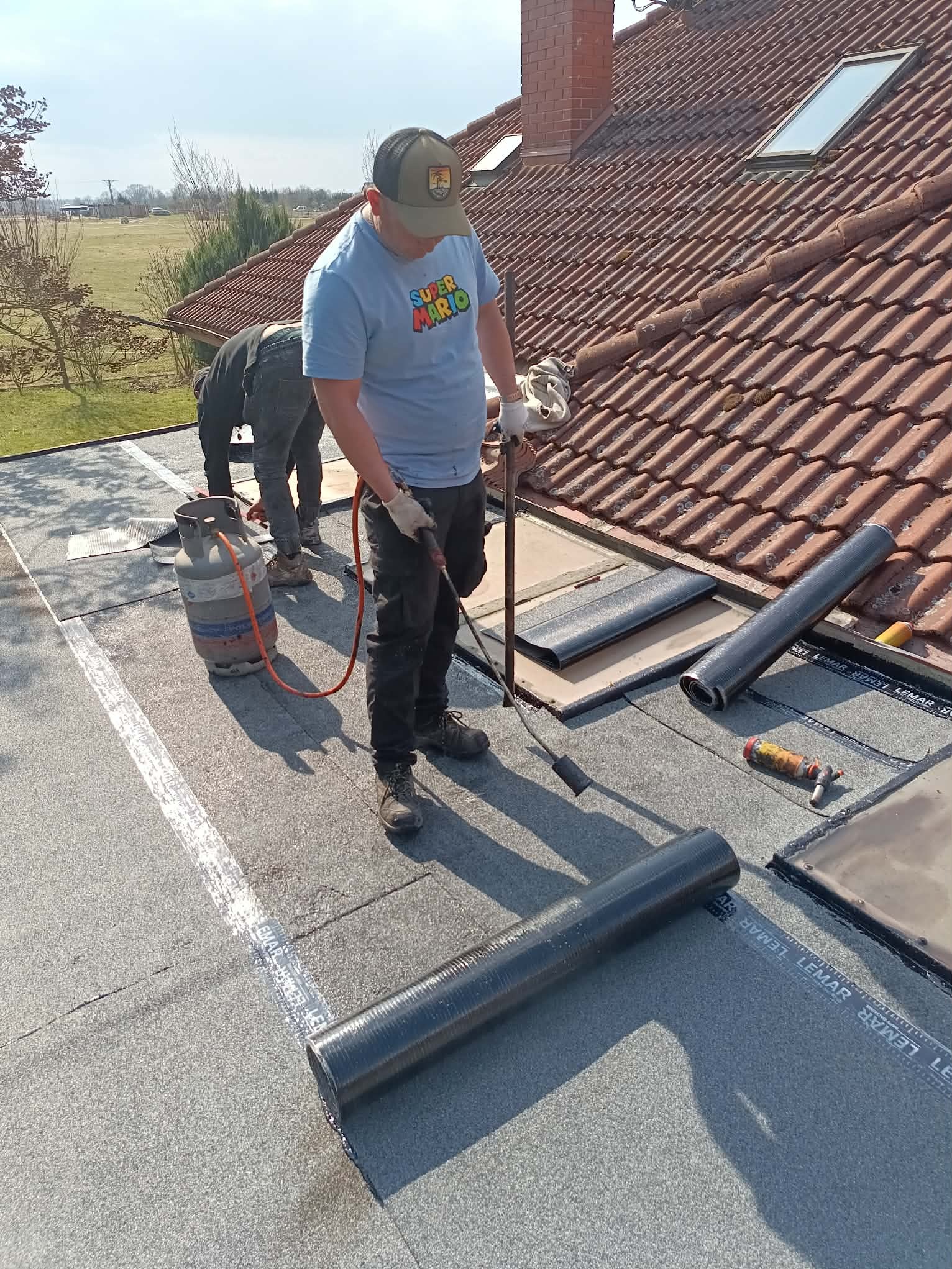 Praca na dachu: dekarz z palnikiem układa papę termozgrzewalną. Widoczna butla gazowa, narzędzia i fragment dachu z oknem. Słoneczny dzień.