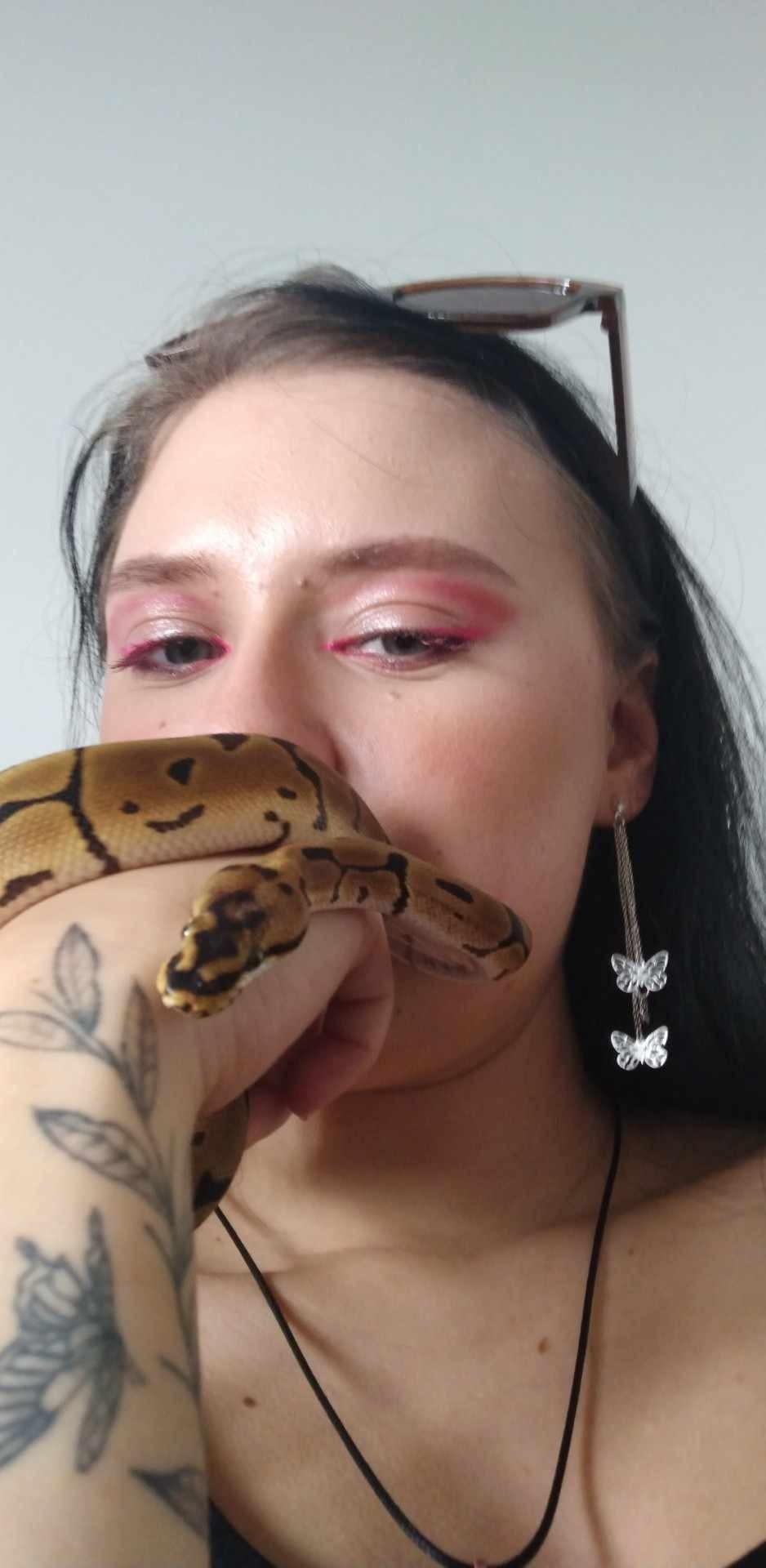 Kobieta z różowym makijażem oczu trzyma na ręce pytona królewskiego, widoczne kolczyki z motylami i tatuaż liści na przedramieniu.