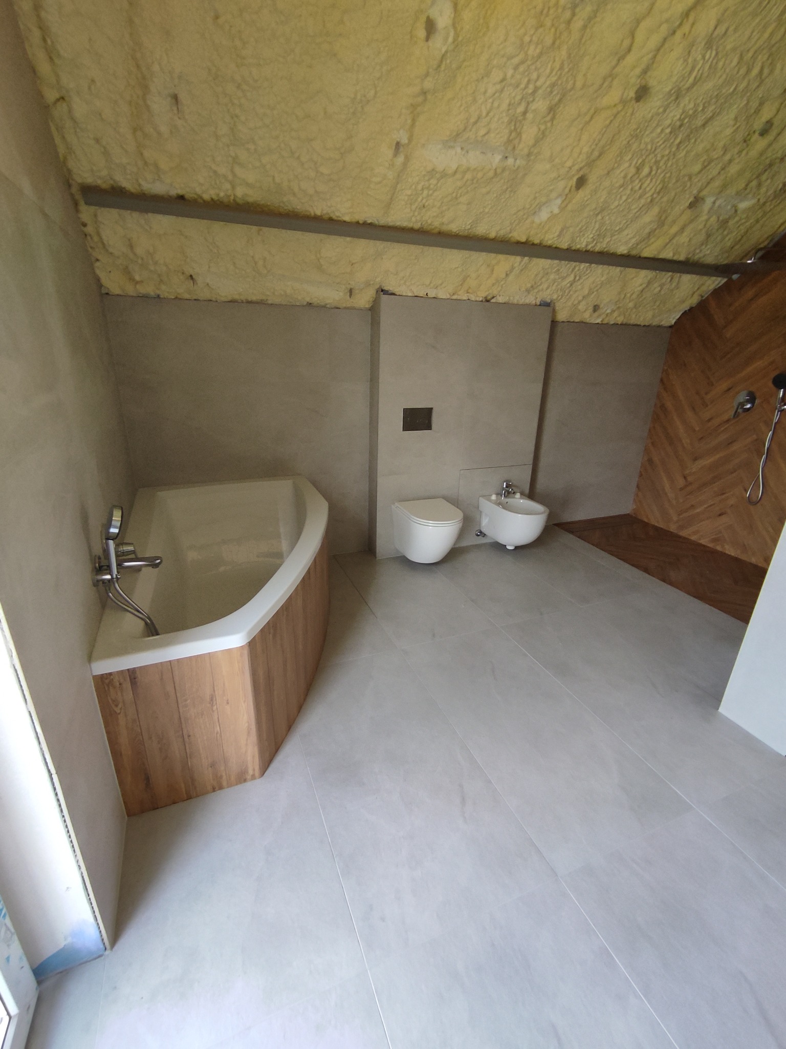 Wnętrze łazienki w trakcie prac wykończeniowych na poddaszu: widoczna wanna z obudową imitującą drewno, toaleta, bidet, prysznic z drewnianą mozaiką ułożoną w jodełkę oraz ocieplenie pianką...