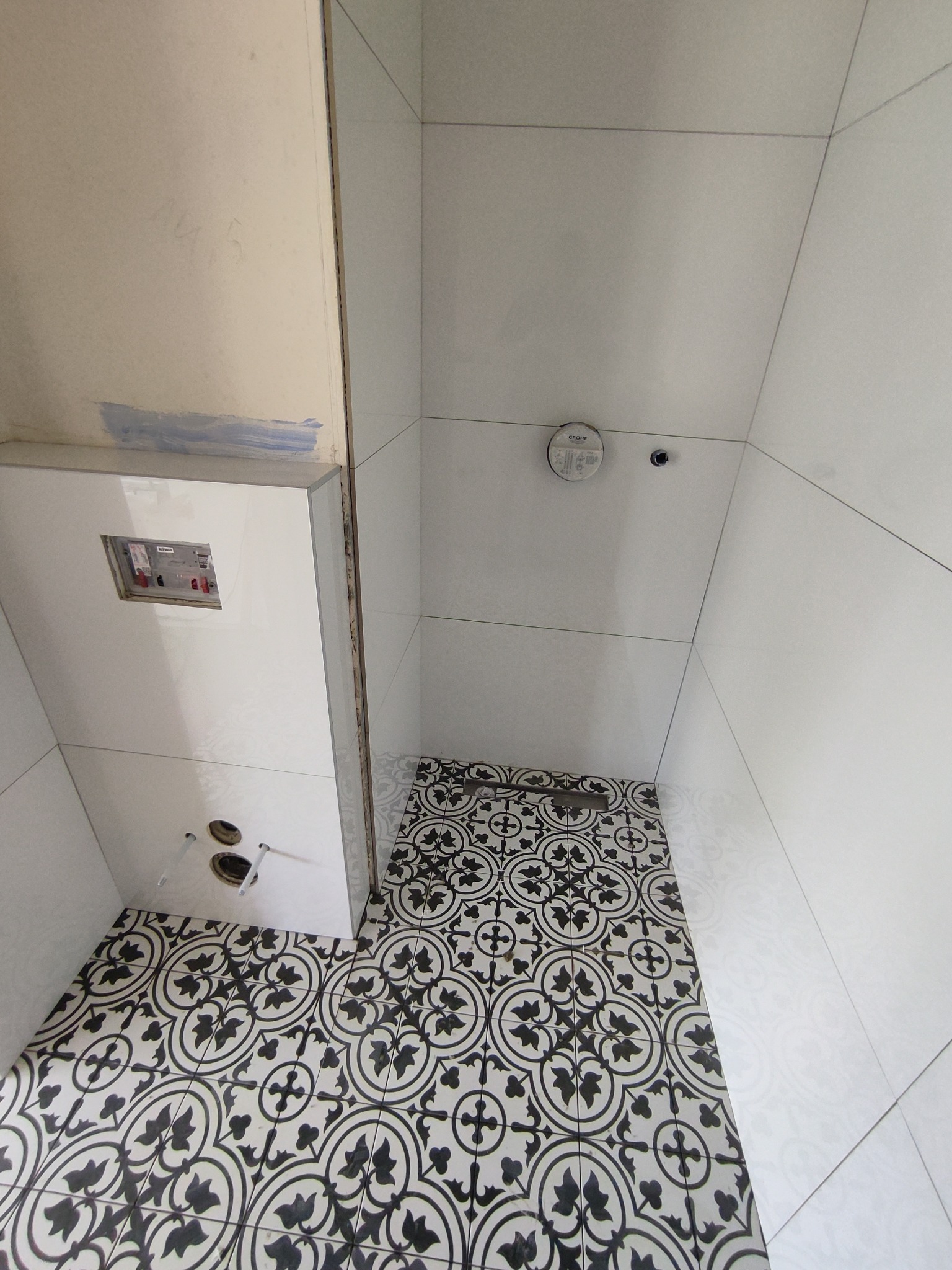 Wnętrze łazienki w trakcie prac wykończeniowych: białe płytki ścienne, dekoracyjna podłoga w czarno-białe wzory, obudowa stelaża WC z widocznymi otworami na instalacje.