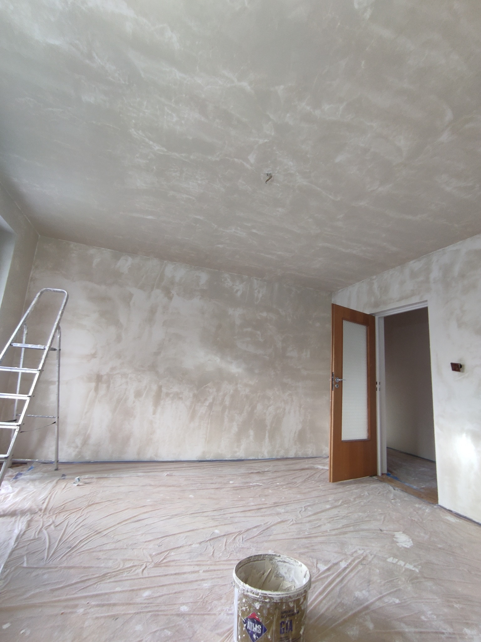 Pokój w trakcie prac wykończeniowych: ściany i sufit pokryte dekoracyjną, nieregularną fakturą, zabezpieczona folią podłoga, aluminiowa drabina oparta o ścianę, wiadro z resztkami farby w rogu.