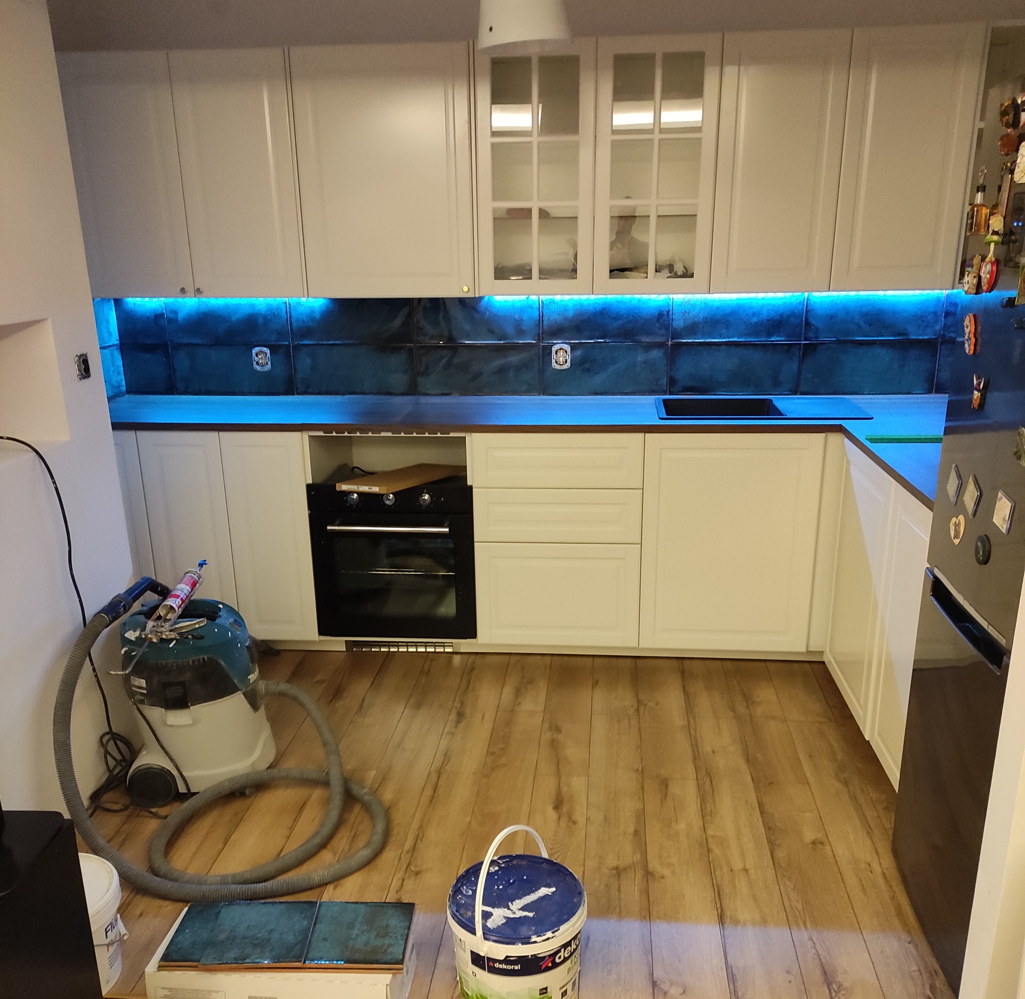 Nowoczesna kuchnia z białymi szafkami, ciemnym blatem i niebieskim oświetleniem LED nad blatem, widoczny odkurzacz i wiadro z farbą, sugerujące prace wykończeniowe.