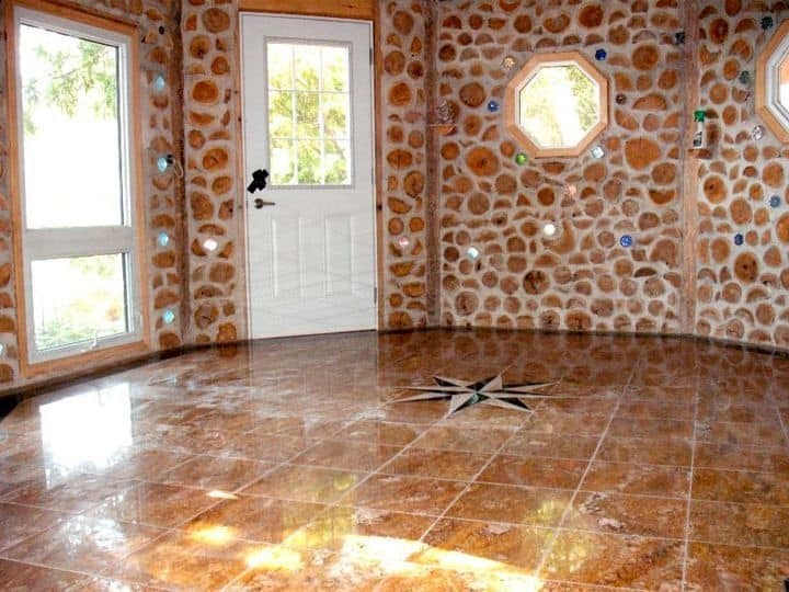 Wnętrze okrągłego pokoju z ścianami wyłożonymi drewnianymi krążkami i kolorowymi szkiełkami, błyszcząca podłoga z motywem gwiazdy w centrum, okna i białe drzwi.