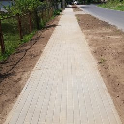 FIRMA USŁUGOWA EWELINA KAGANIUK, TOMASZ KOWALCZYK SPÓŁKA CYWILNA - Nowo ułożony chodnik z jasnoszarej kostki brukowej wzdłuż drogi, z widocznym przygotowanym podłożem po bokach. W tle zieleń, dom i przechodnie.