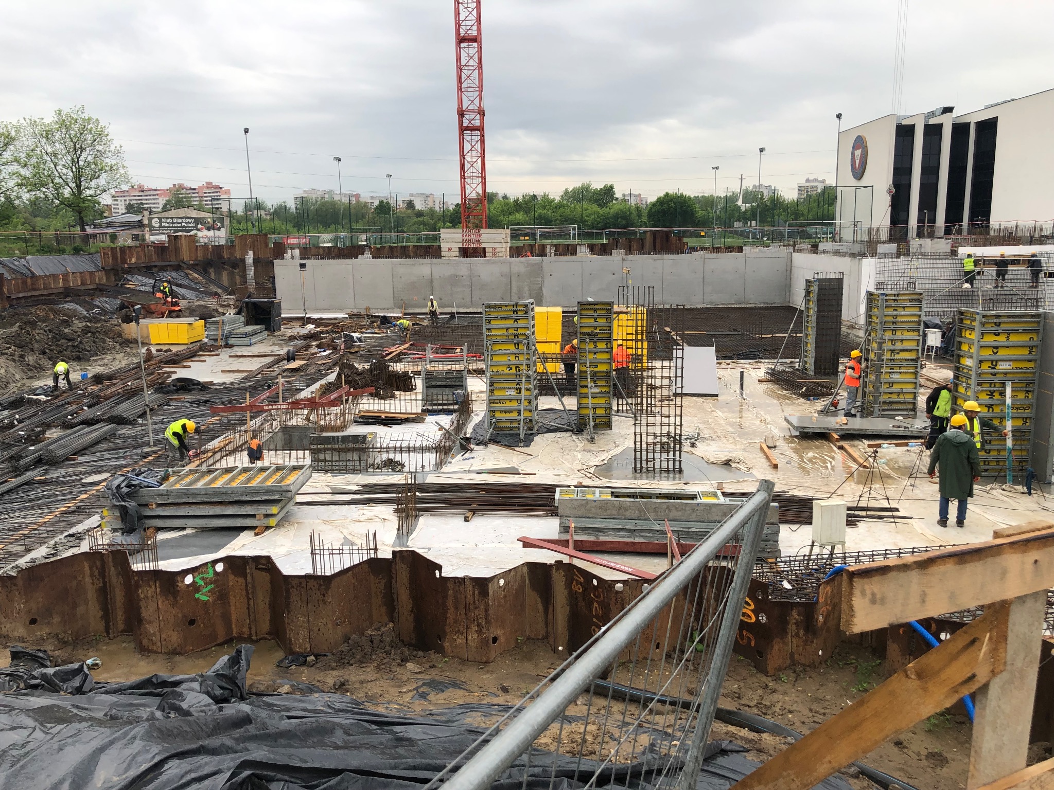 Szeroki kadr placu budowy z widocznymi zbrojeniami, szalunkami w kolorze żółtym, pracownikami w kaskach i elementami konstrukcyjnymi fundamentów, z budynkiem w tle i żurawiem na horyzoncie.