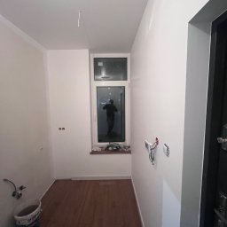 Wykończenia wnętrz - Wnętrze pomieszczenia w trakcie remontu: widoczne drewniane panele podłogowe, białe ściany z odkrytymi przewodami elektrycznymi, okno z widokiem na zewnątrz i wiadro farby w rogu.