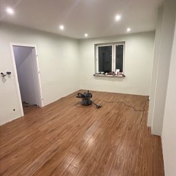 Wykończenia wnętrz - Pokój po remoncie z nową drewnianą podłogą, widoczna maszyna do polerowania podłóg, okno i drzwi w tle, oświetlenie sufitowe.