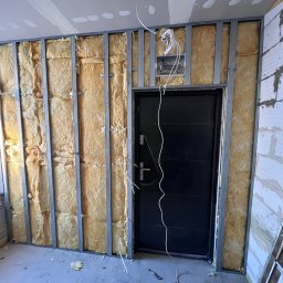 Wykończenia wnętrz - Wnętrze w trakcie prac: montaż metalowej konstrukcji z izolacją z wełny mineralnej przy ścianie z pustaków i czarnych drzwiach, widoczne przewody elektryczne i drabina.