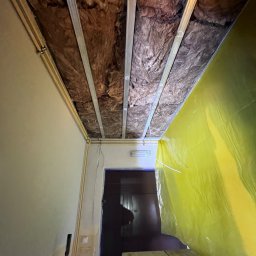 Wykończenia wnętrz - Widok z dołu na sufit w trakcie montażu, widoczna izolacja z wełny mineralnej, metalowy stelaż oraz żółta folia ochronna, zabezpieczająca przed pyłem.