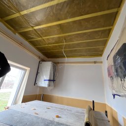 Wykończenia wnętrz - Przygotowanie do prac malarskich w pomieszczeniu: sufit zabezpieczony folią malarską, grzejnik i urządzenia elektryczne osłonięte folią, widoczny stół roboczy z resztkami materiałów malarskich.