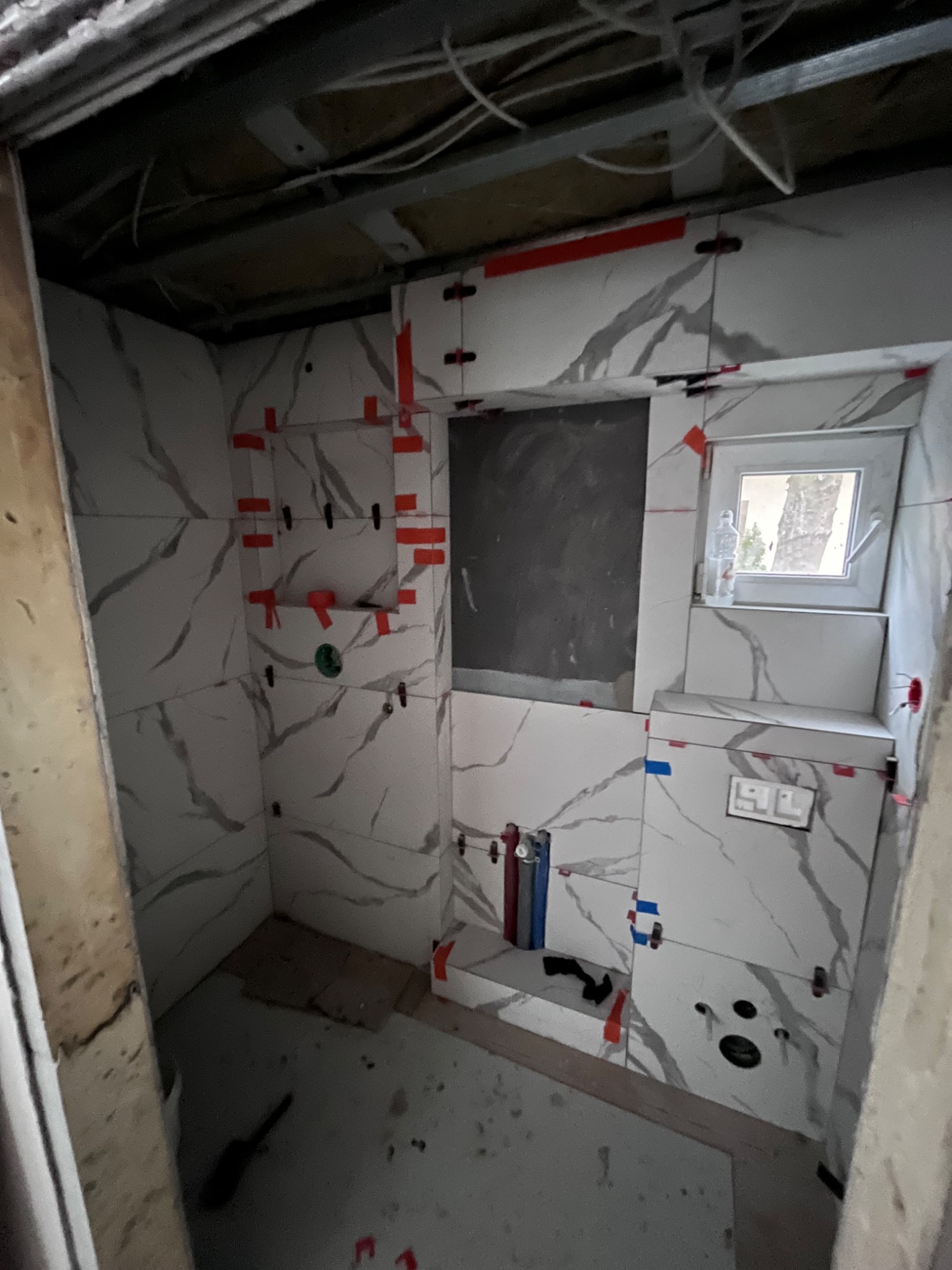 Ściany łazienki w trakcie kładzenia płytek imitujących marmur, widoczne plastikowe krzyżyki dystansowe, niezabudowana wnęka okienna z butelką wody na parapecie, oraz wystające rury instalacji wodnej.