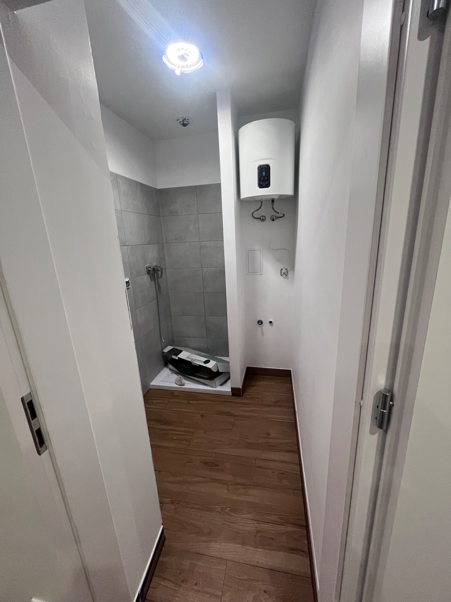 Wnętrze małej łazienki w trakcie prac wykończeniowych, widoczny bojler na ścianie, prysznic z szarą glazurą, podłoga z paneli imitujących drewno oraz karton po akcesoriach prysznicowych na podłodze.