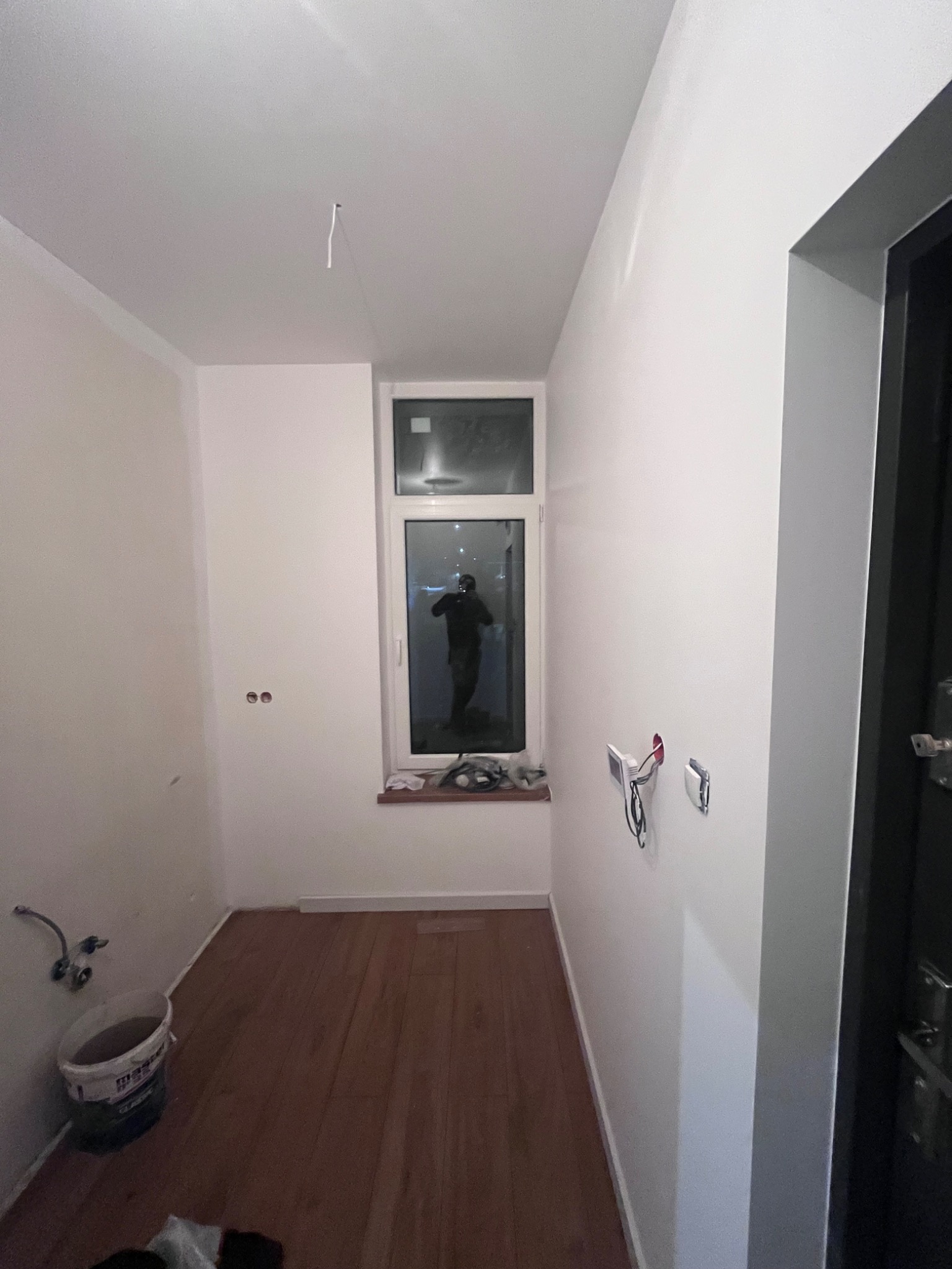 Wnętrze pomieszczenia w trakcie remontu: widoczne drewniane panele podłogowe, białe ściany z odkrytymi przewodami elektrycznymi, okno z widokiem na zewnątrz i wiadro farby w rogu.