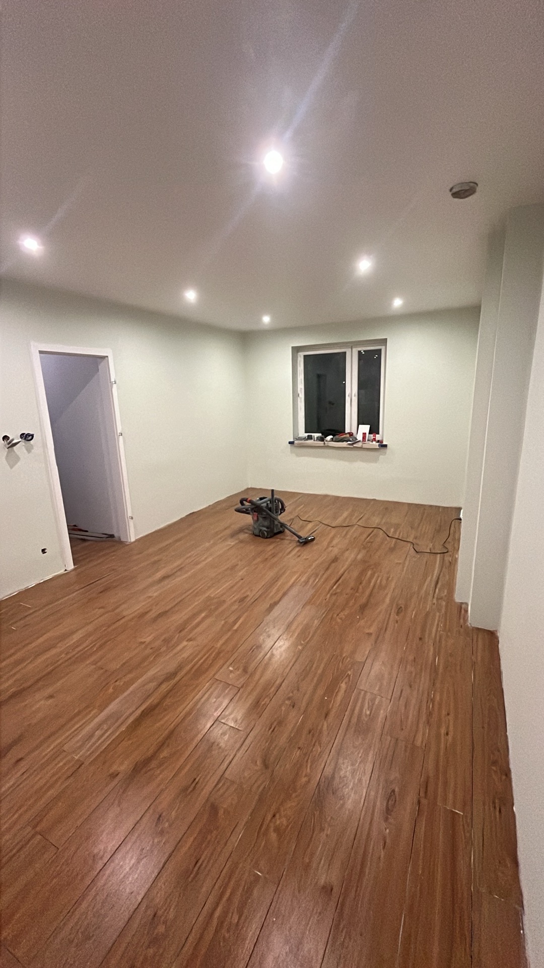 Pokój po remoncie z nową drewnianą podłogą, widoczna maszyna do polerowania podłóg, okno i drzwi w tle, oświetlenie sufitowe.