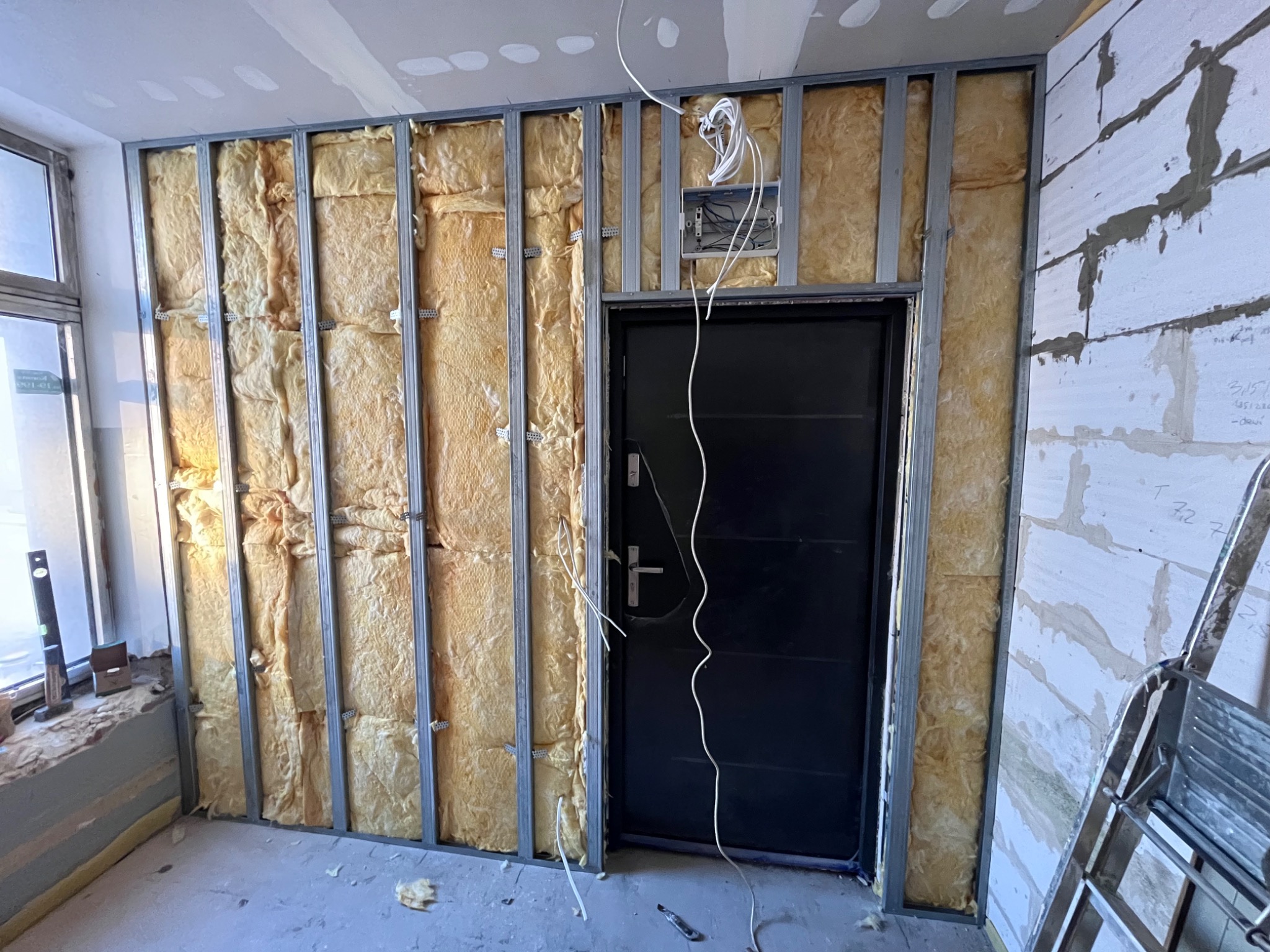 Wnętrze w trakcie prac: montaż metalowej konstrukcji z izolacją z wełny mineralnej przy ścianie z pustaków i czarnych drzwiach, widoczne przewody elektryczne i drabina.