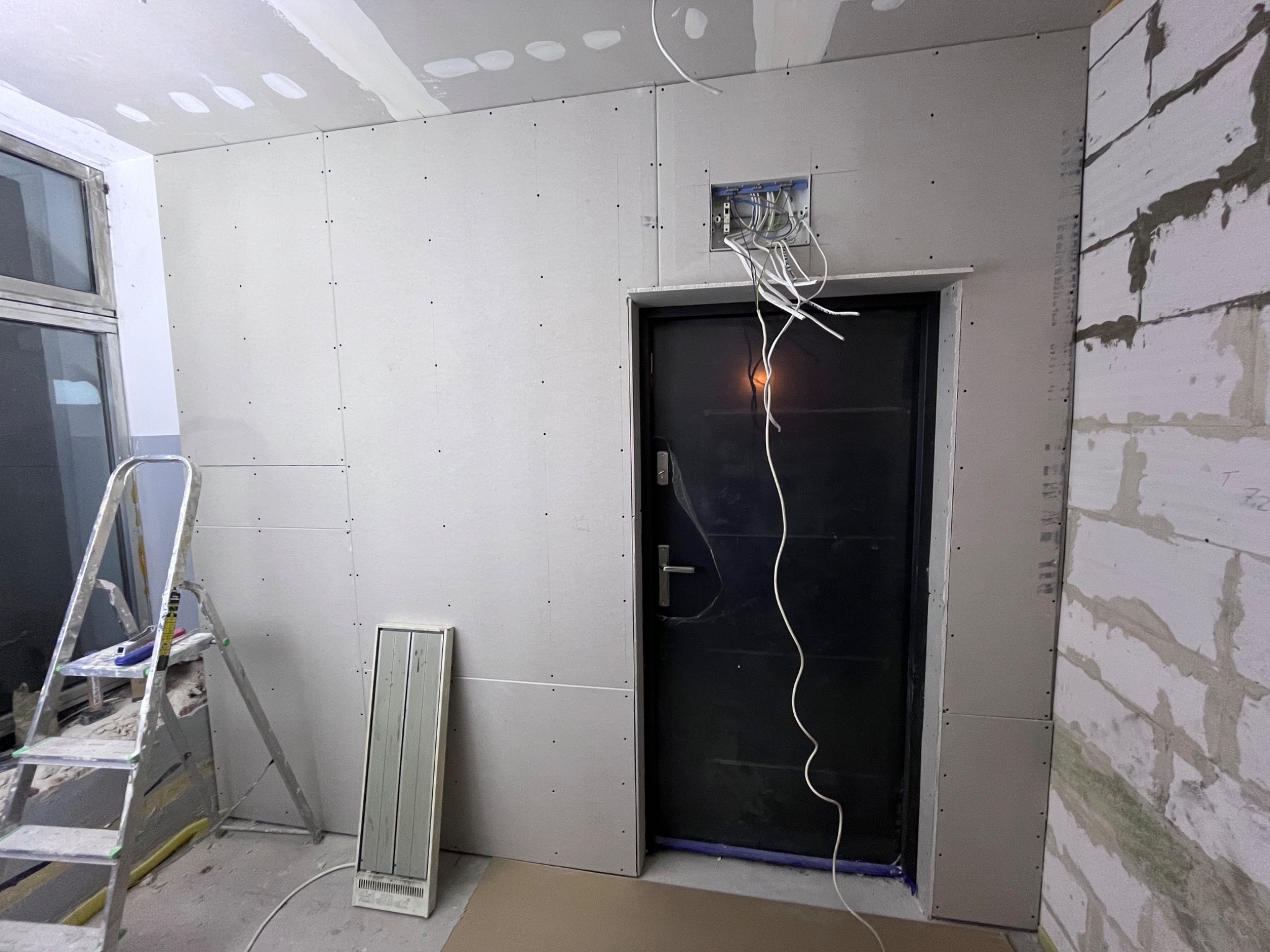 Wnętrze w trakcie remontu: ściany obłożone płytami gipsowo-kartonowymi, widoczna skrzynka elektryczna z wystającymi przewodami nad ciemnymi drzwiami, drabina i grzejnik przenośny w pomieszczeniu.