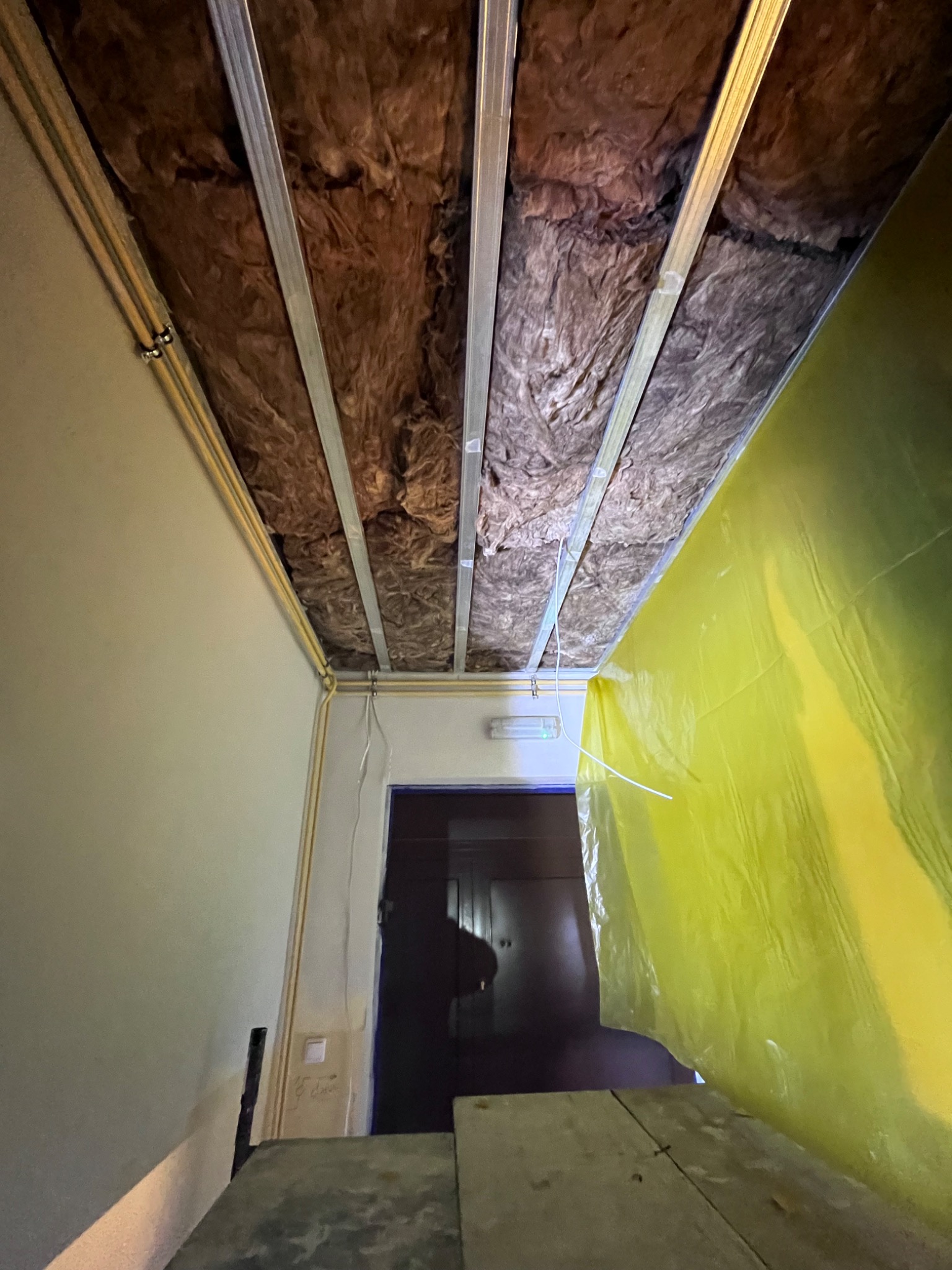 Widok z dołu na sufit w trakcie montażu, widoczna izolacja z wełny mineralnej, metalowy stelaż oraz żółta folia ochronna, zabezpieczająca przed pyłem.