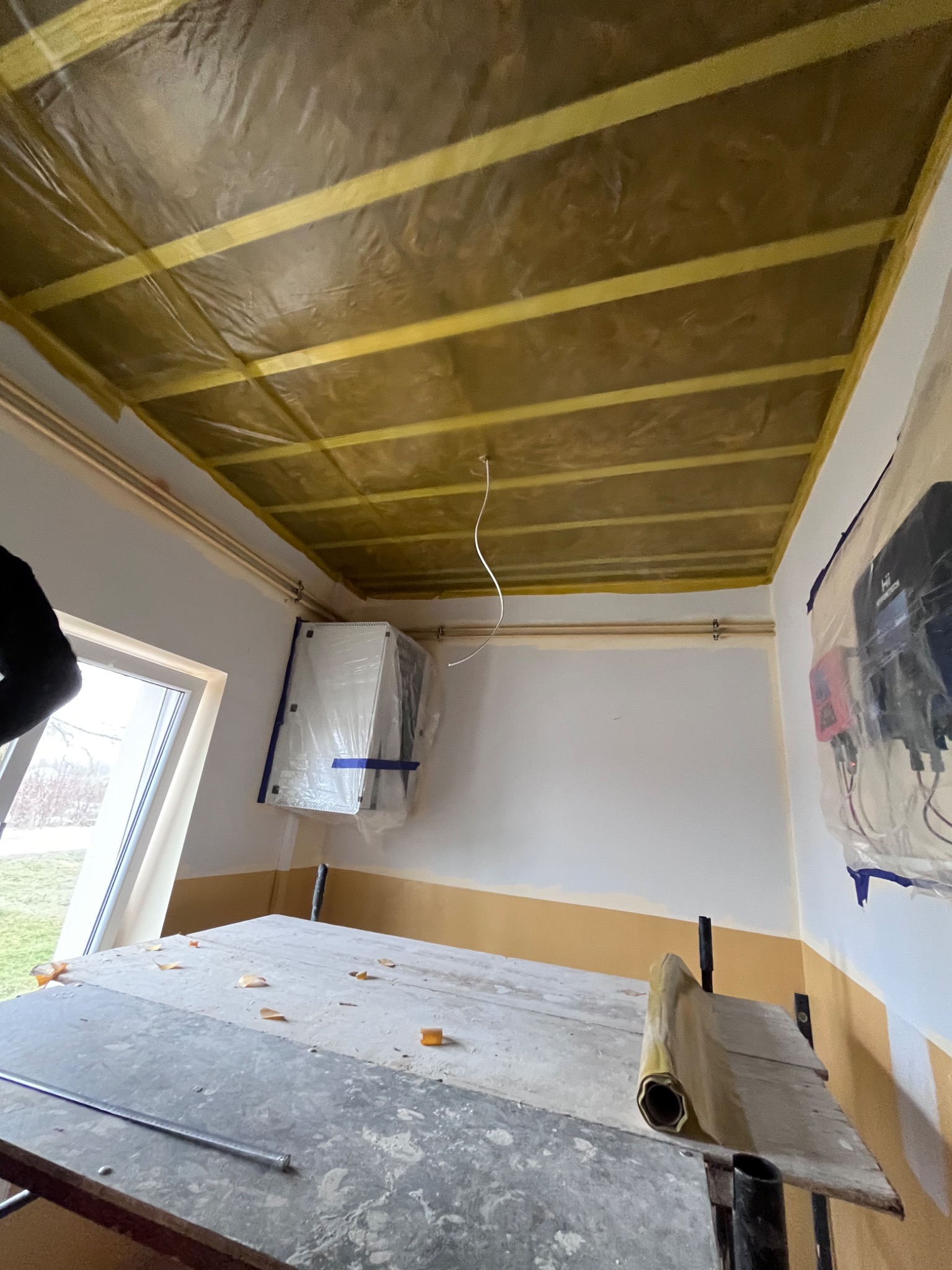 Przygotowanie do prac malarskich w pomieszczeniu: sufit zabezpieczony folią malarską, grzejnik i urządzenia elektryczne osłonięte folią, widoczny stół roboczy z resztkami materiałów malarskich.