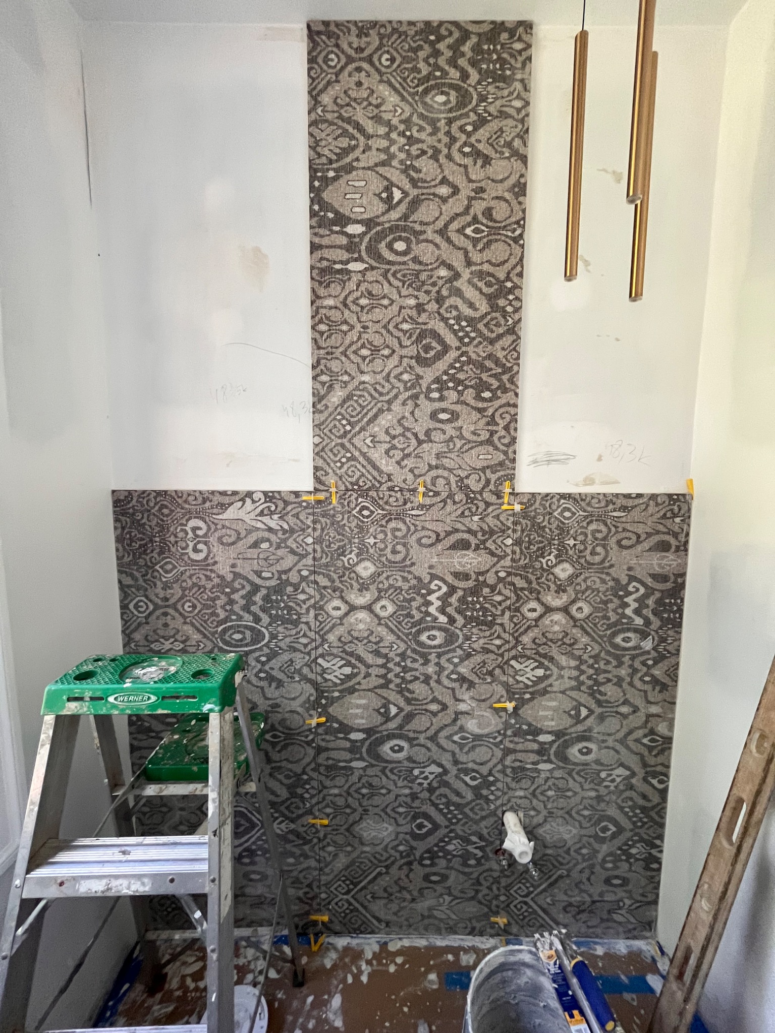Montaż płytek z wzorem etnicznym w łazience, widoczna drabina, poziomica i krzyżyki dystansowe, surowe ściany powyżej płytek, wiszące lampy.