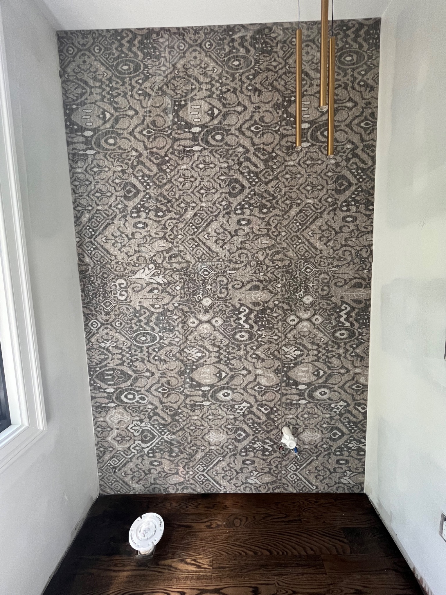 Fragment pokoju z tapetą w etniczne wzory, drewnianą podłogą i złotymi lampami wiszącymi, widoczne niedokończone prace wykończeniowe przy ścianach.