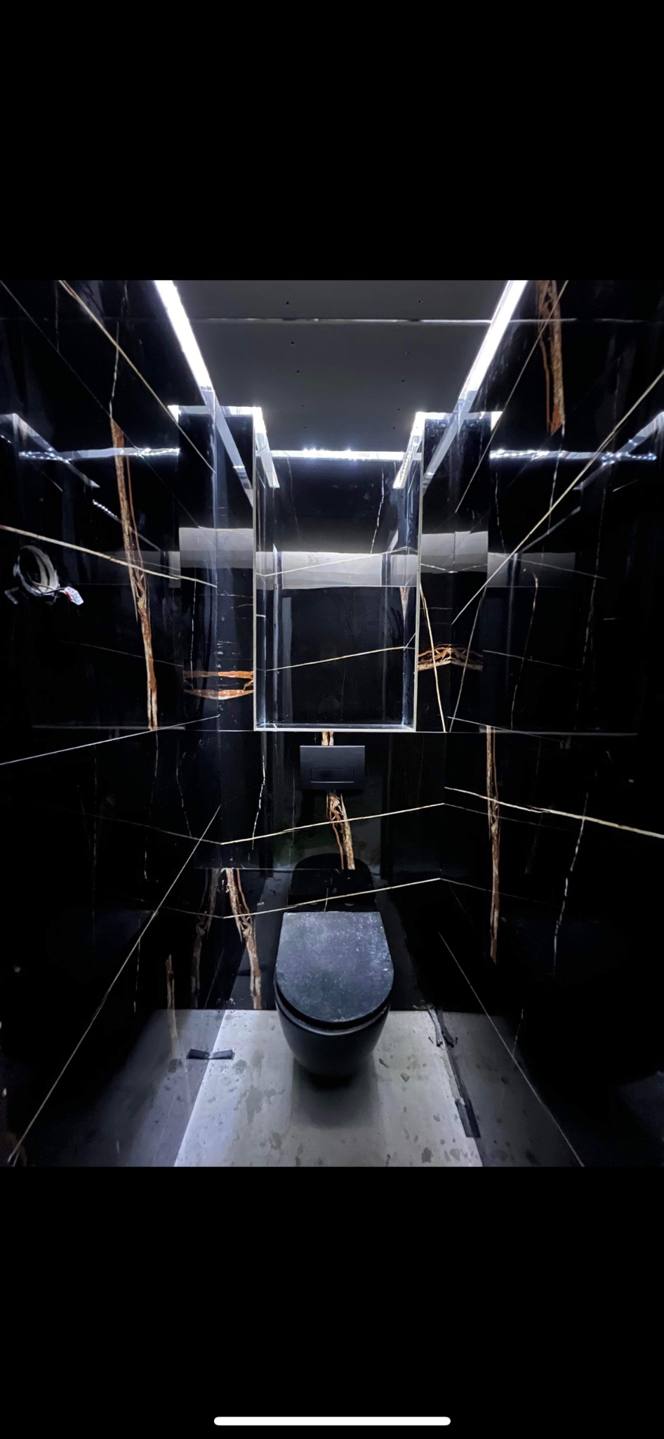 Czarne płytki imitujące marmur z złotymi żyłami w małej toalecie, podświetlane lustro, wisząca muszla klozetowa.