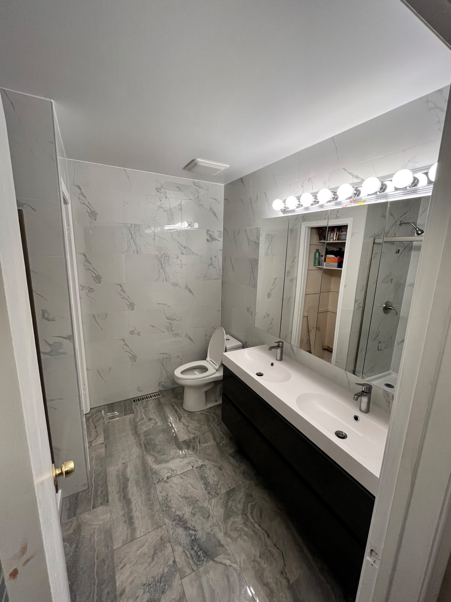 Wykończona łazienka z marmurowymi płytkami ściennymi i podłogowymi, białą umywalką z dwoma kranami na czarnej szafce, toaletą i dużym lustrem z oświetleniem punktowym.