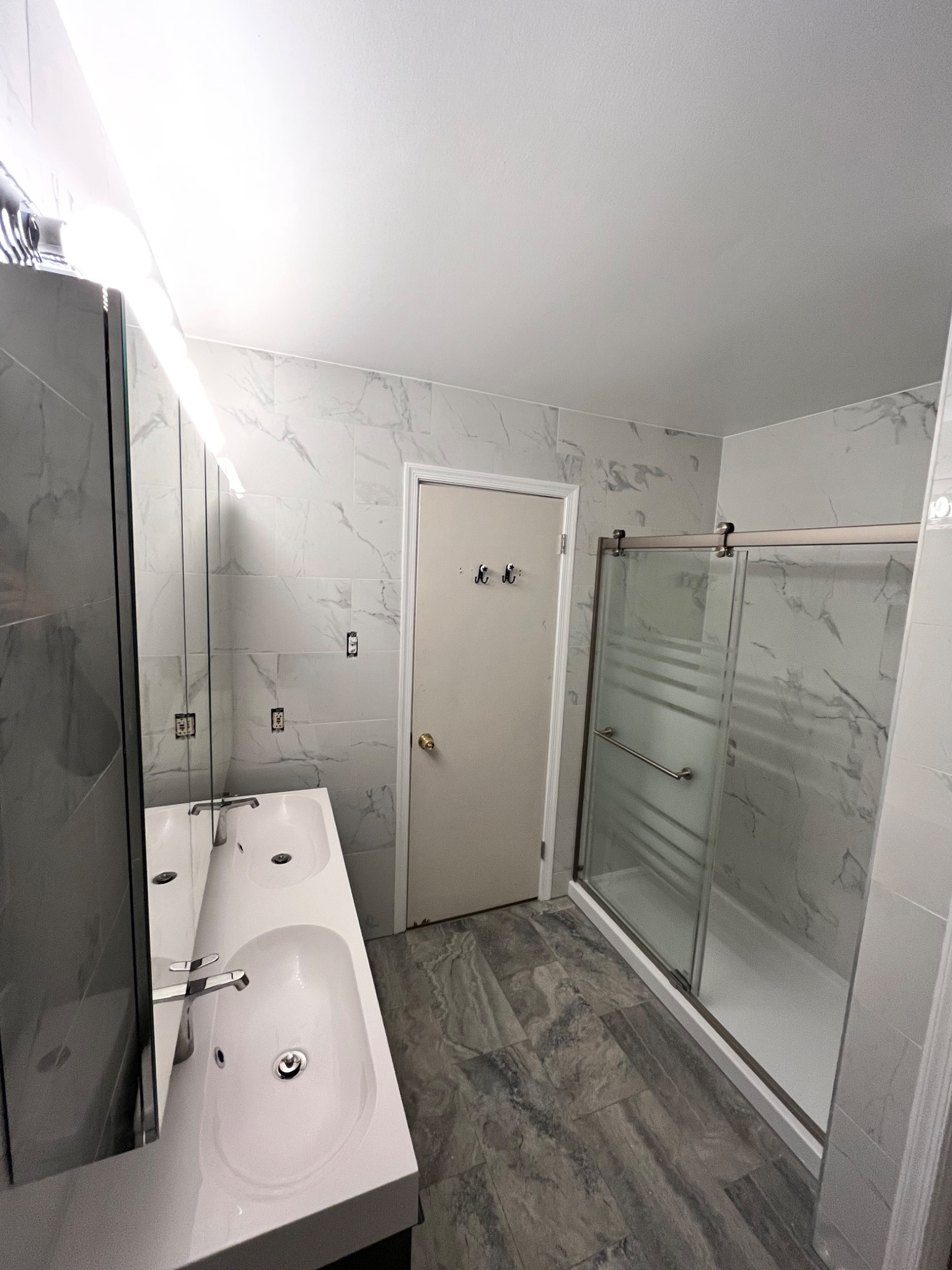 Wyremontowana łazienka z podwójną umywalką nablatową, kabiną prysznicową ze szklanymi przesuwnymi drzwiami i płytkami imitującymi marmur na ścianach oraz podłogą z paneli winylowych w odcieniach...