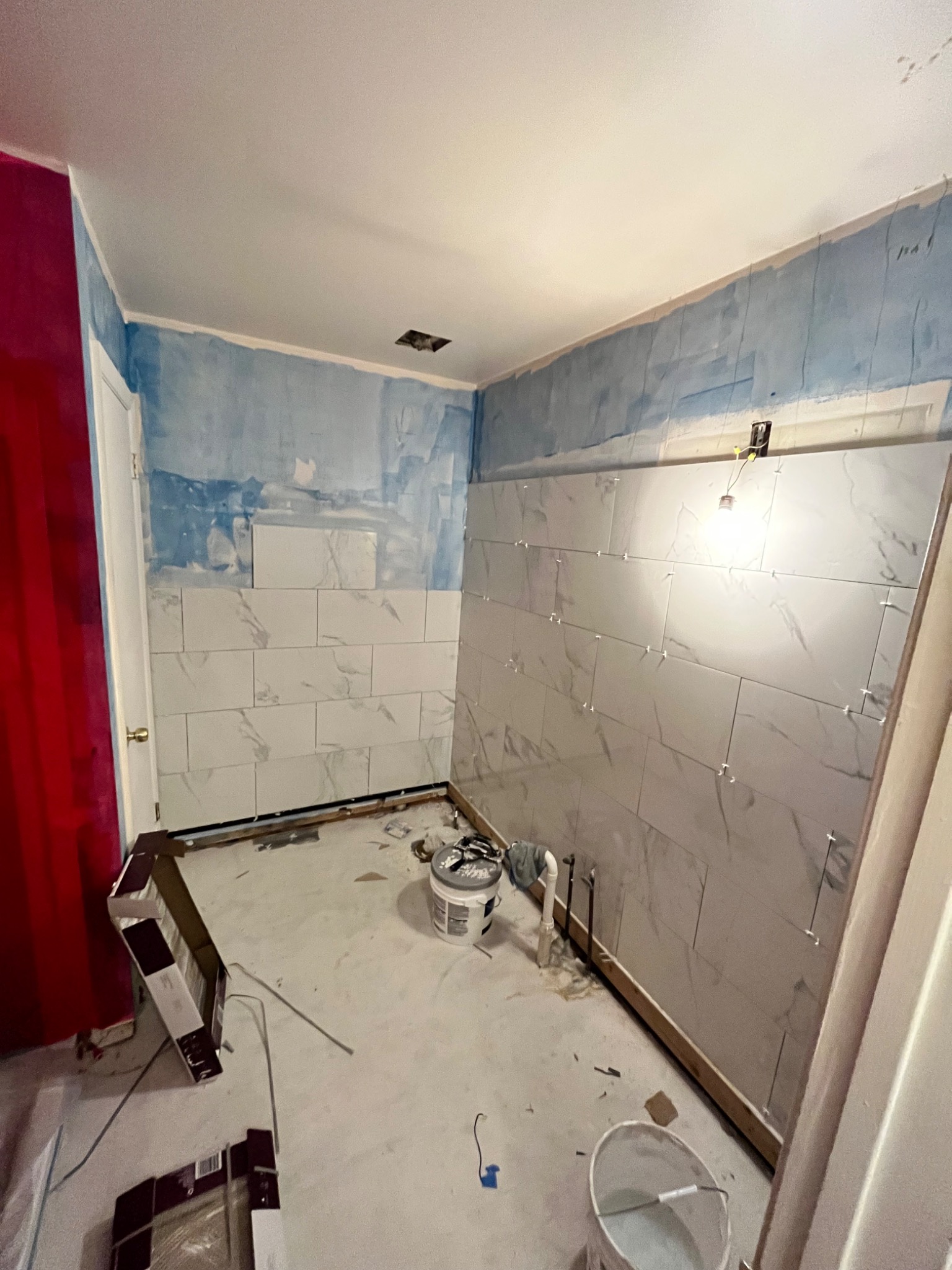 Remont łazienki w trakcie: częściowo wyłożona ściana płytkami imitującymi marmur, widoczne krzyżyki dystansowe, niebieska farba podkładowa na ścianach, rury instalacyjne, kartony po płytkach...