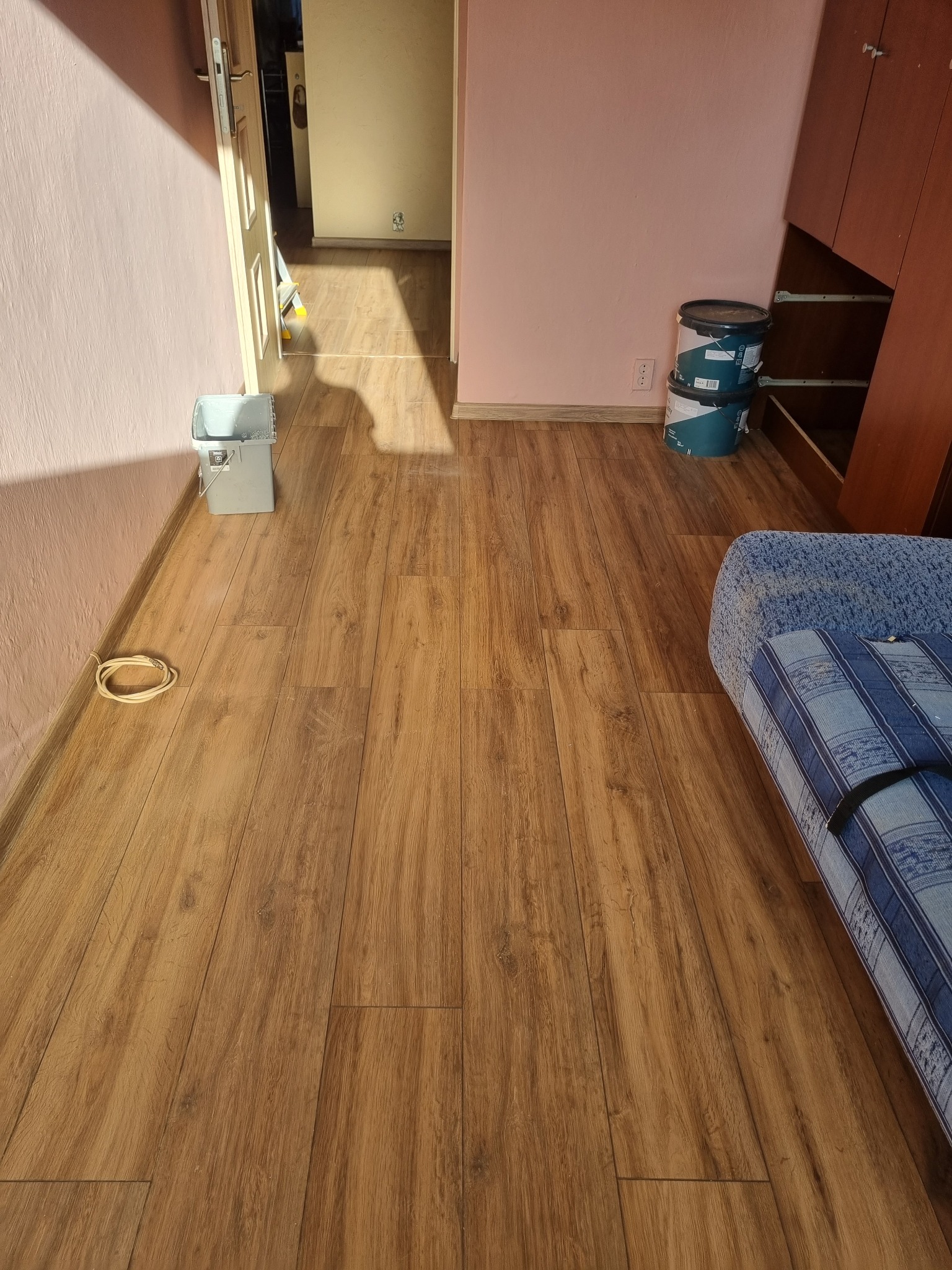 Podłoga z paneli w mieszkaniu w Warszawie, widoczne wiadro z farbą i puszki z farbą przy ścianie, częściowo zasłonięta sofa.