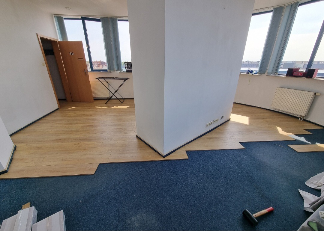 Proces układania paneli podłogowych w jasnym pomieszczeniu biurowym, widoczne fragmenty ułożonej podłogi, niebieska wykładzina dywanowa, młotek i paczka paneli w dolnym rogu.