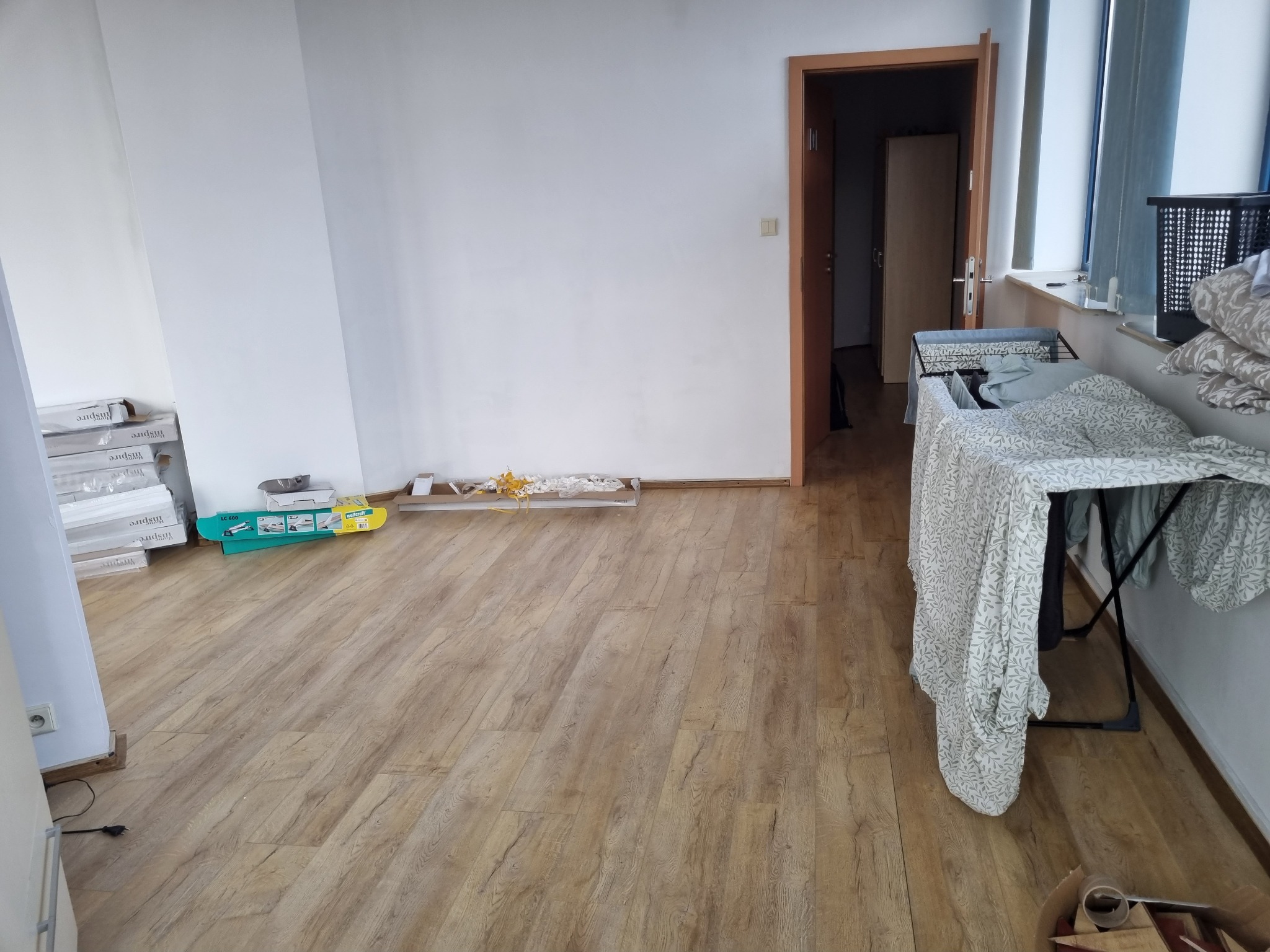 Pokój w trakcie remontu: ułożone panele podłogowe, przy ścianie paczki paneli i narzędzia, w tle widoczne drzwi do innego pomieszczenia.