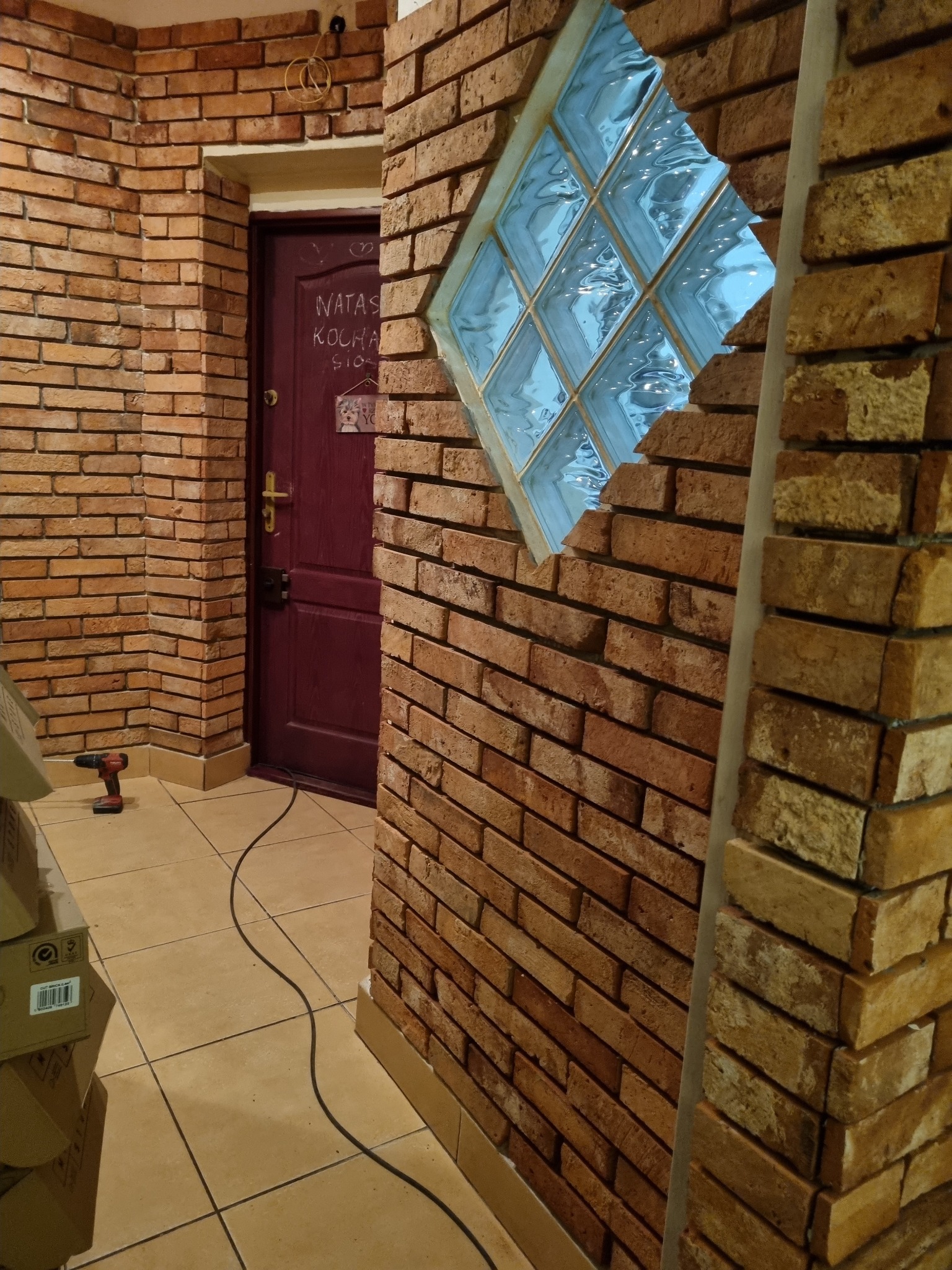 Wnętrze z ceglaną ścianą, w której zamontowano luksfery w kształcie rombu, widoczny fragment podłogi z beżowych płytek, w tle bordowe drzwi z napisem, w rogu wiertarka akumulatorowa i kartony.