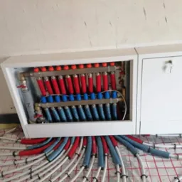 Rozdzielacz ogrzewania podłogowego w białej szafce z kluczykiem, rury grzewcze czerwone i niebieskie, widoczne mocowania do podłoża.