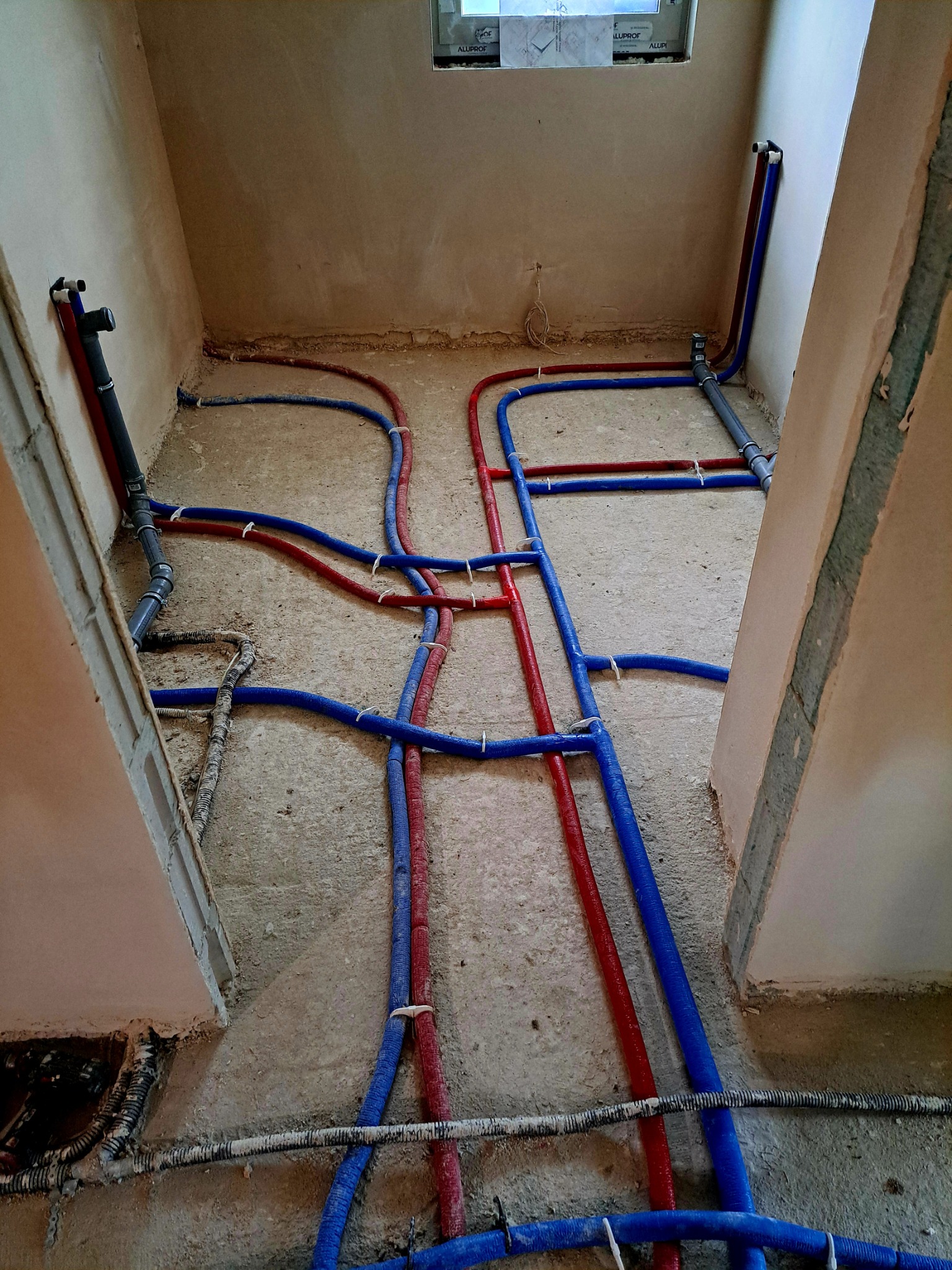 Nowa instalacja hydrauliczna i ogrzewania podłogowego w surowym stanie, rury PEX w kolorze czerwonym i niebieskim, mocowane do podłoża w pomieszczeniu z oknem.