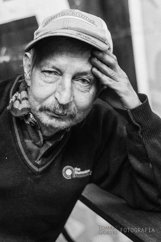 Czarno-biały portret starszego mężczyzny w czapce z daszkiem, zamyślony wyraz twarzy, dłoń oparta o czoło, widoczne logo na swetrze.