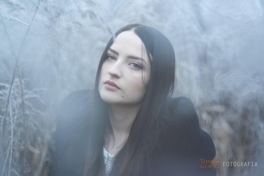 Portret kobiety z ciemnymi włosami, spoglądającej w górę, na tle zarośli pokrytych szronem, z efektem rozmycia w dolnej części kadru.