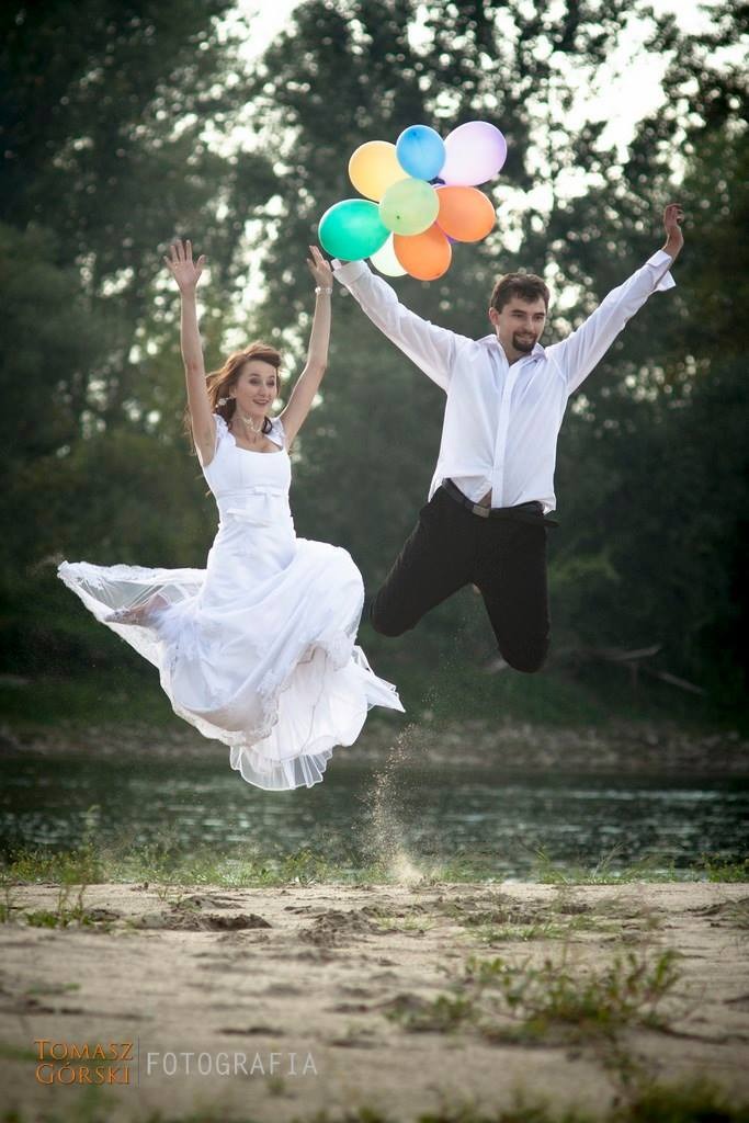 Uśmiechnięta para młoda w skoku na plaży, trzymająca kolorowe balony, z piaskiem unoszącym się spod stóp.